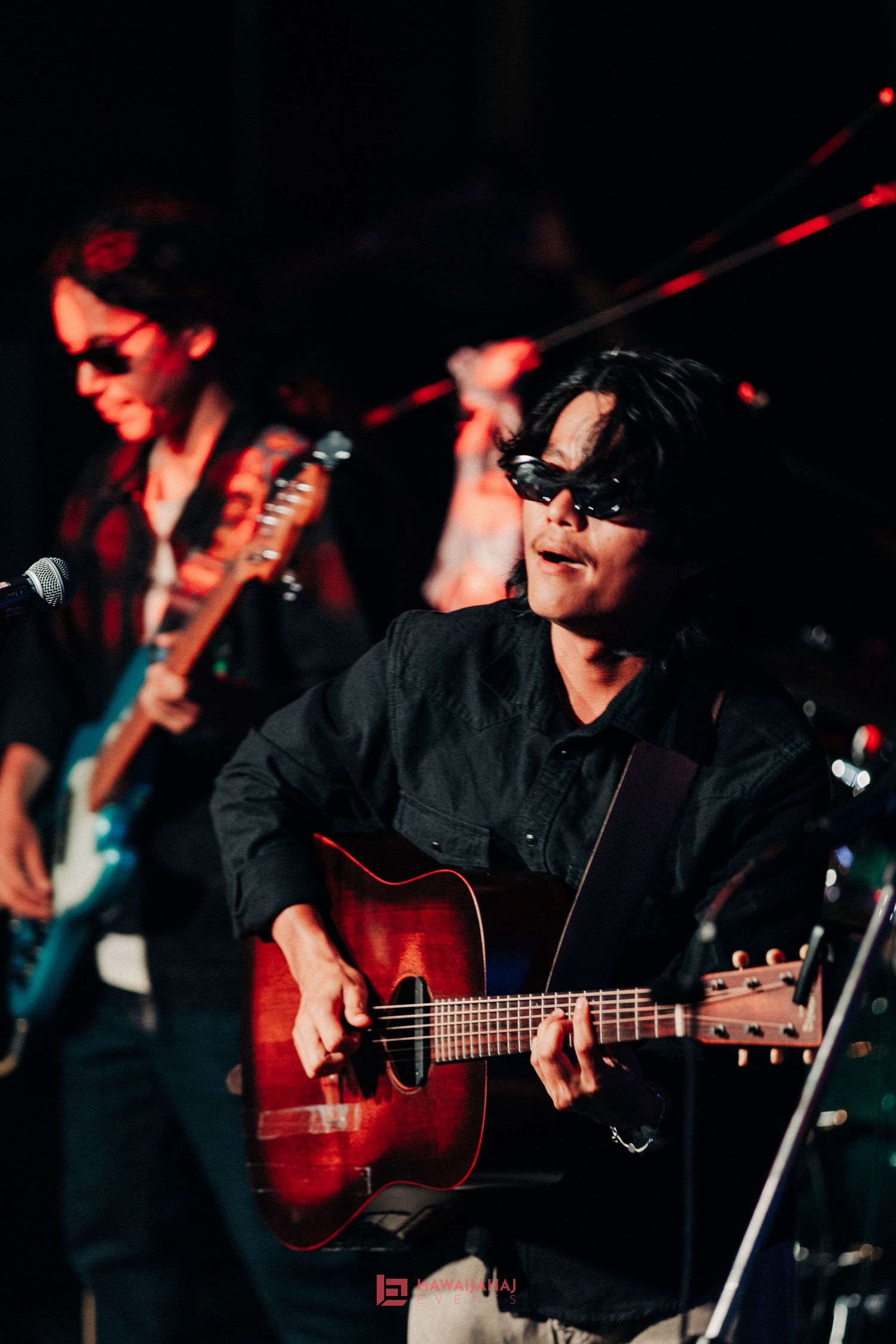 Musicians perform on stage with acoustic guitars in dramatic red lighting during a live concert performance.