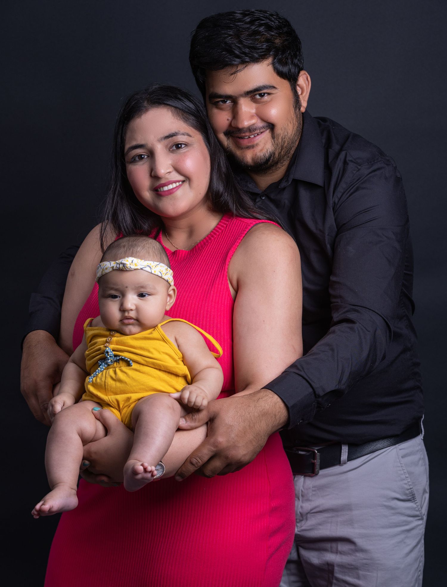 Family portrait with baby in yellow outfit and parent in red against black background.