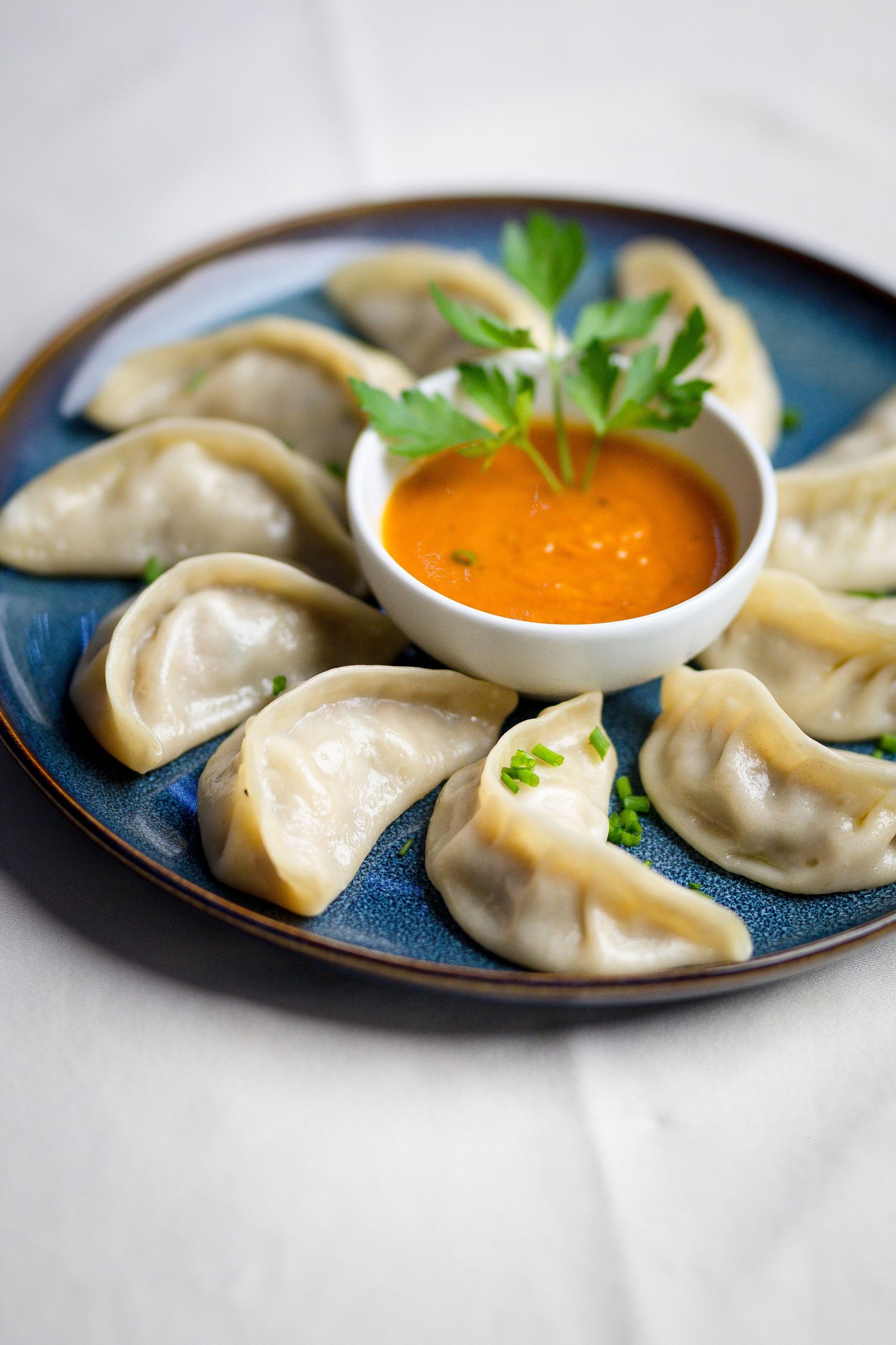 Steamed dumplings arranged on blue plate with orange dipping sauce and fresh herb garnish.