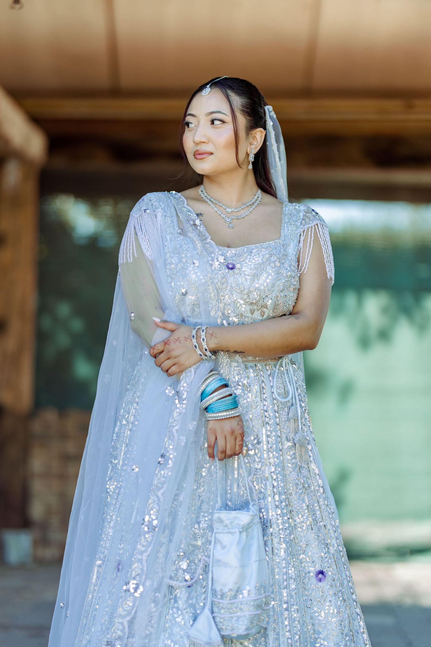 Bride in shimmering white lehenga with crystal details poses under covered outdoor area.