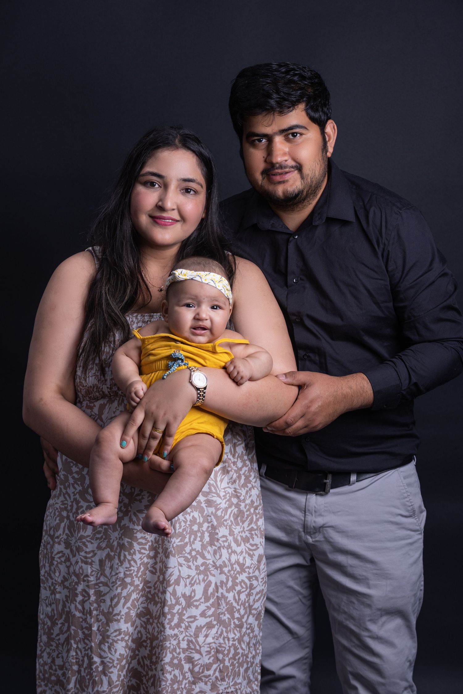 Family poses in formal attire against dark backdrop with baby in yellow outfit.