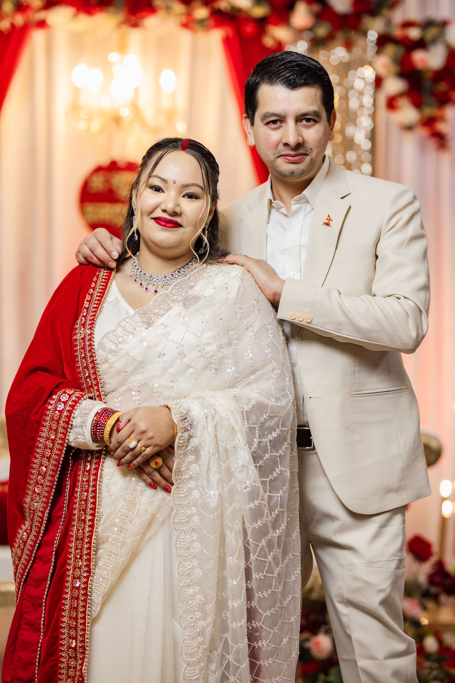 Couple in traditional South Asian formal wear posing against floral decorated backdrop.