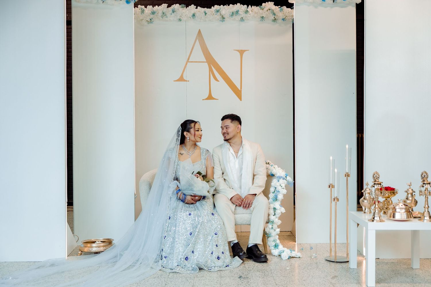 A couple sits together at an elegant wedding ceremony setup with gold monogram letters and white floral decorations.