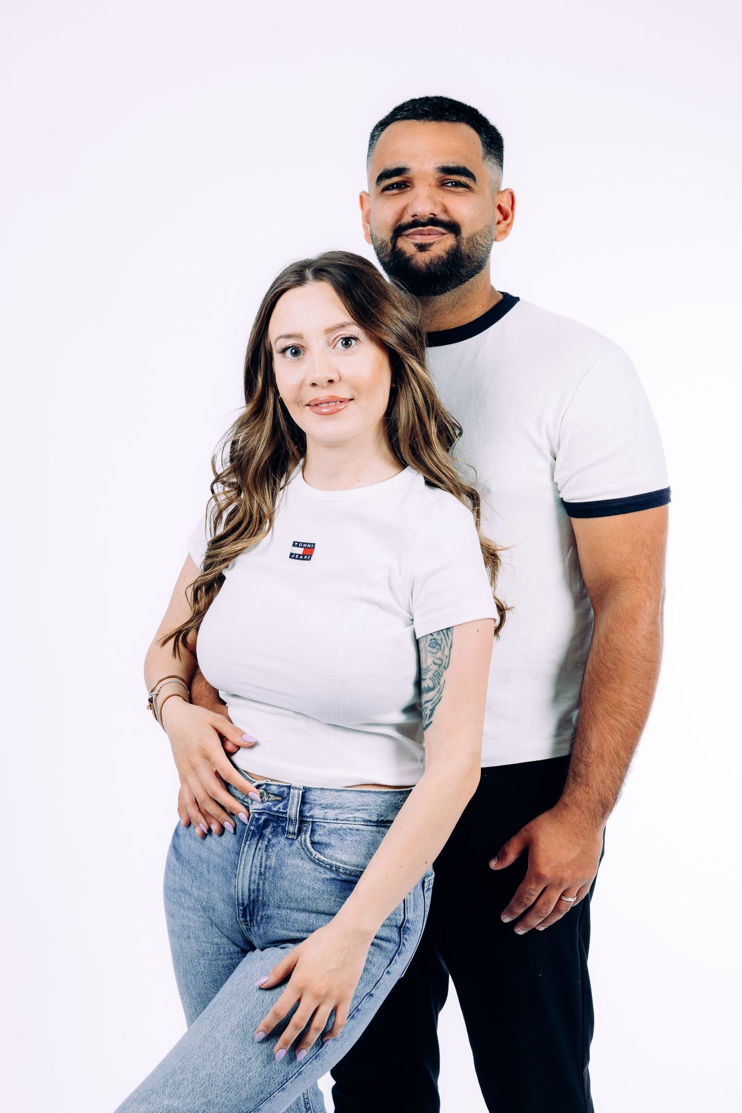 Casual couple in white shirts and jeans posing against white background in studio setting.