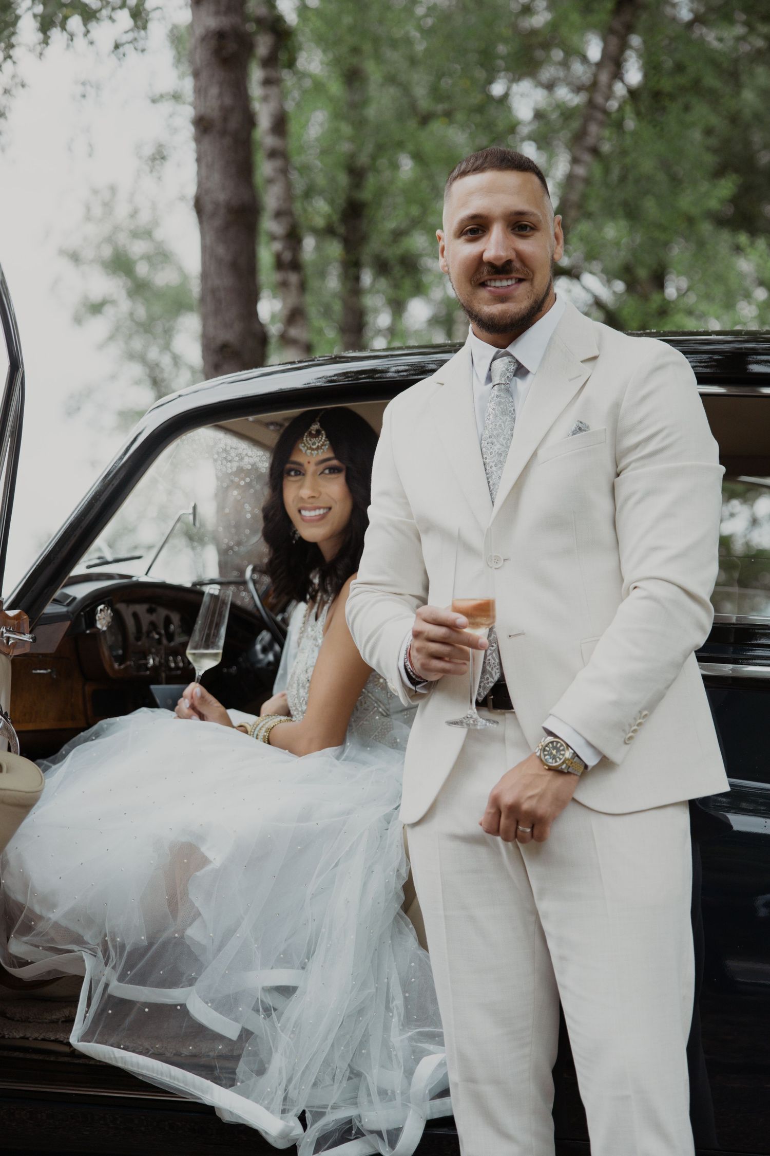 A couple celebrates with champagne beside their vintage black car in a forest setting.