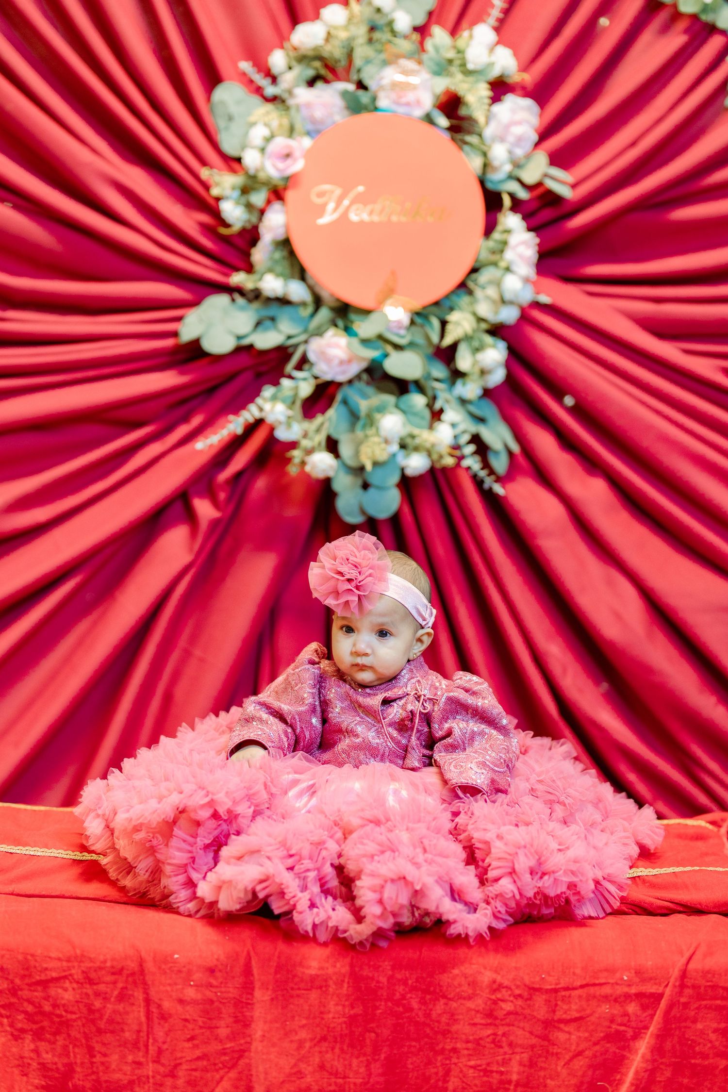Baby wearing pink tulle dress against dramatic red draped backdrop with floral wreath decoration.
