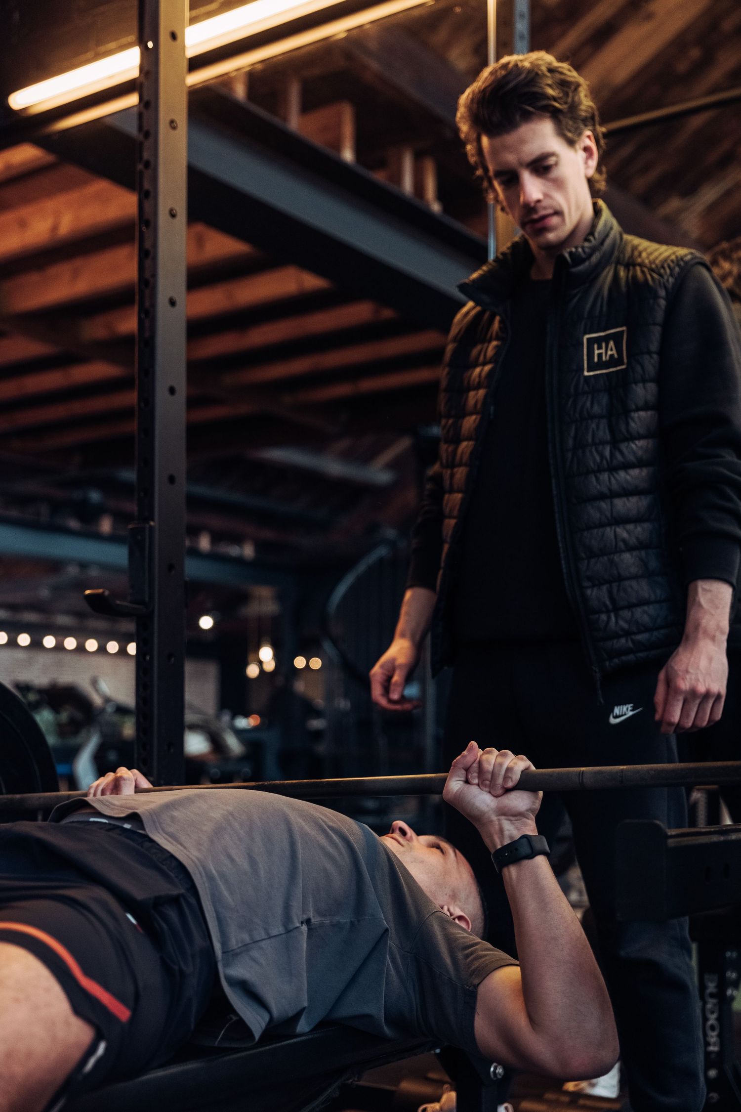 Personal trainer spotting client during bench press workout in dark industrial gym setting.