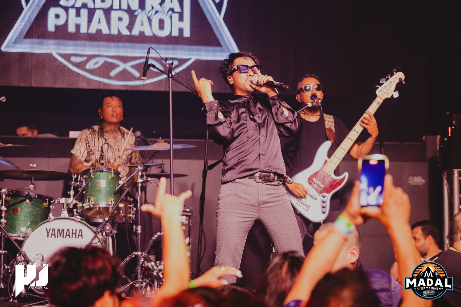 Live band performance with drummer and bassist on stage as crowd takes photos.