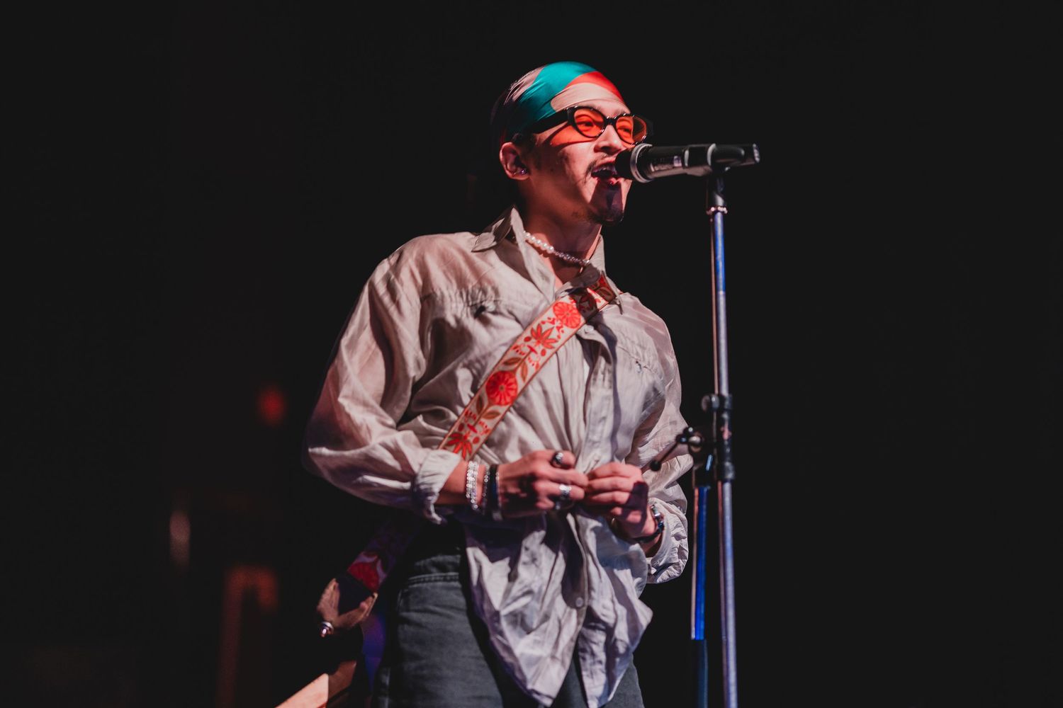 Performer in glasses singing into microphone on darkened stage.