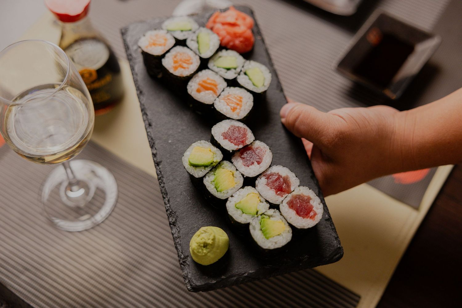 Hands preparing fresh sushi rolls on dark slate board with ingredients.
