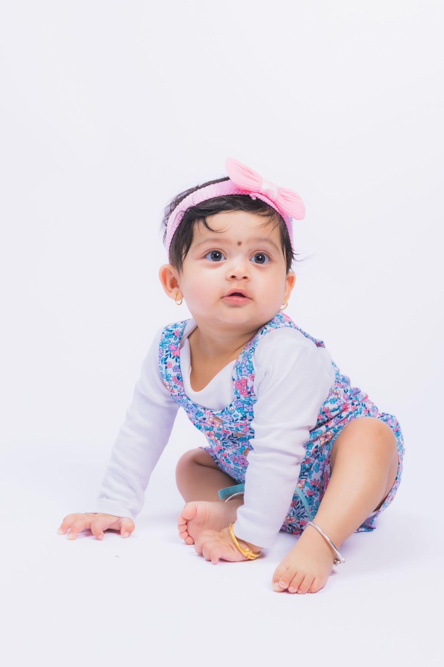 Baby in floral outfit and pink bow crawls on white studio floor.