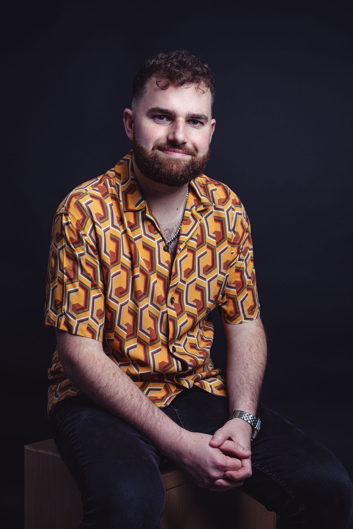 Person wearing geometric pattern shirt poses thoughtfully against dark background.