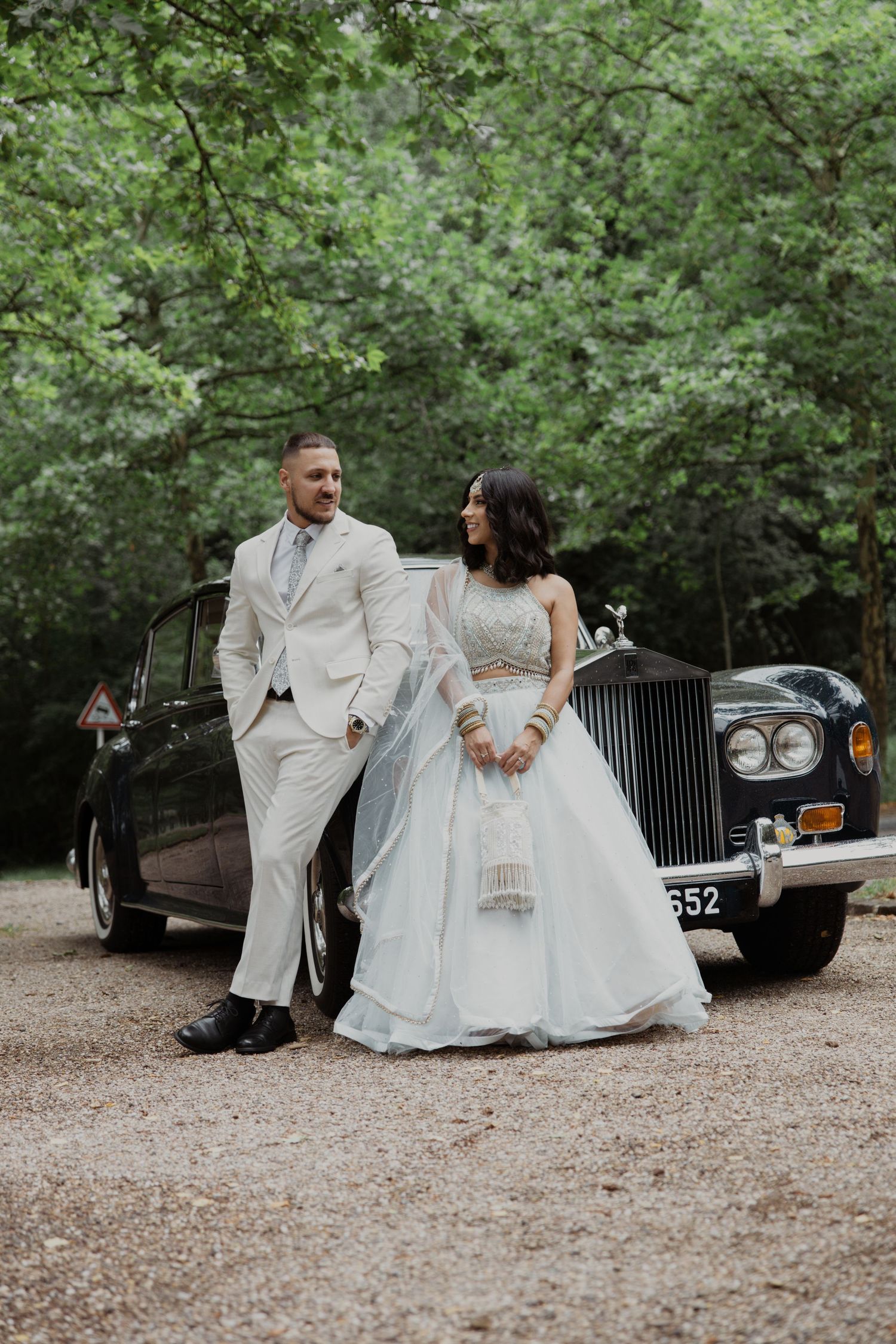 Stylish couple in white wedding attire pose against classic Rolls Royce in wooded setting.