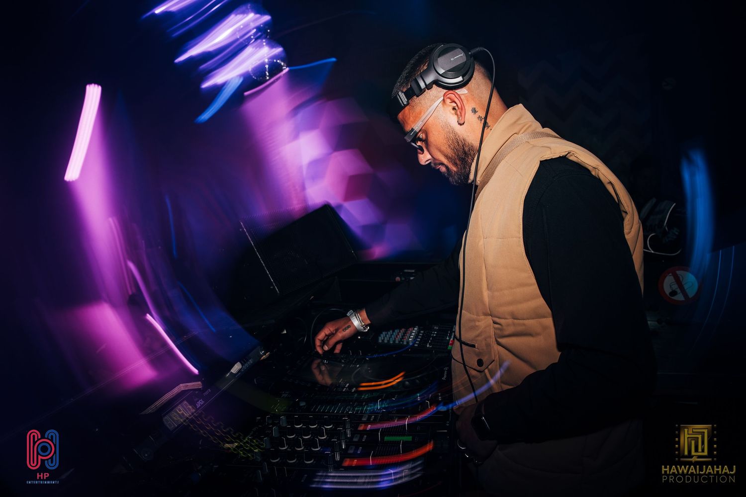 DJ performing at turntables under purple lighting effects wearing vest and sunglasses.