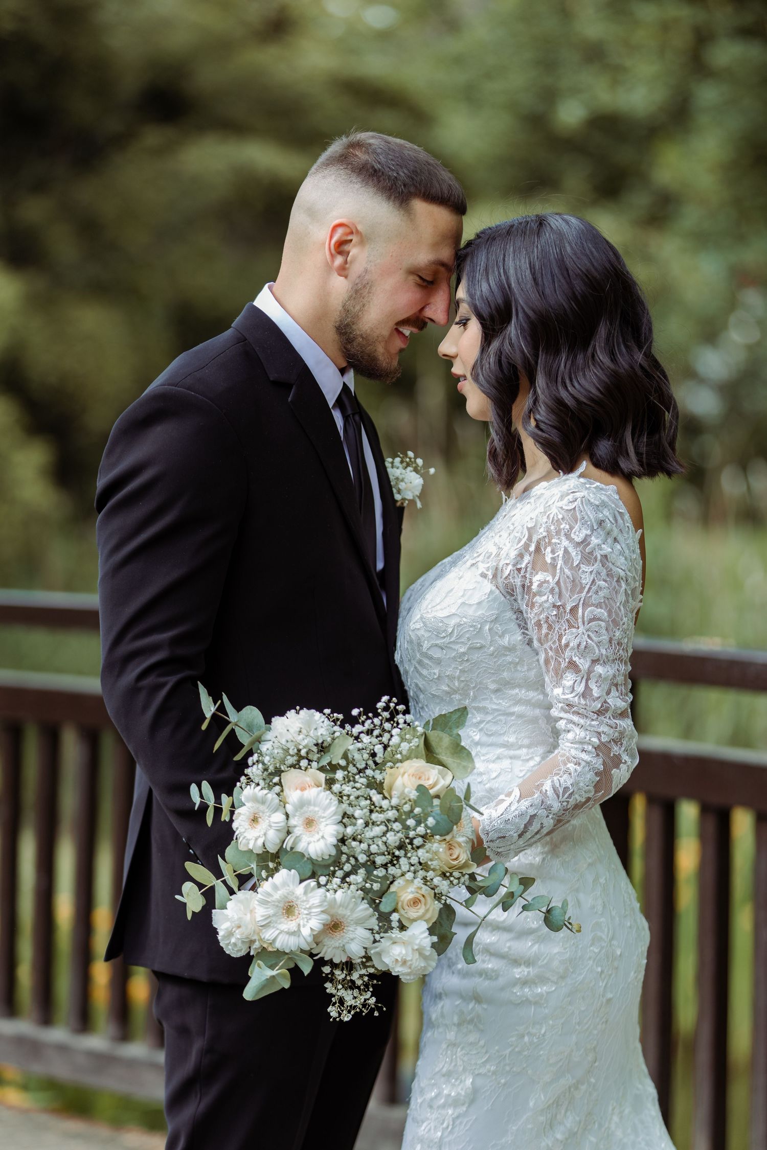 Wedding couple in white lace dress and black suit holding bouquet of white flowers on wooden deck.