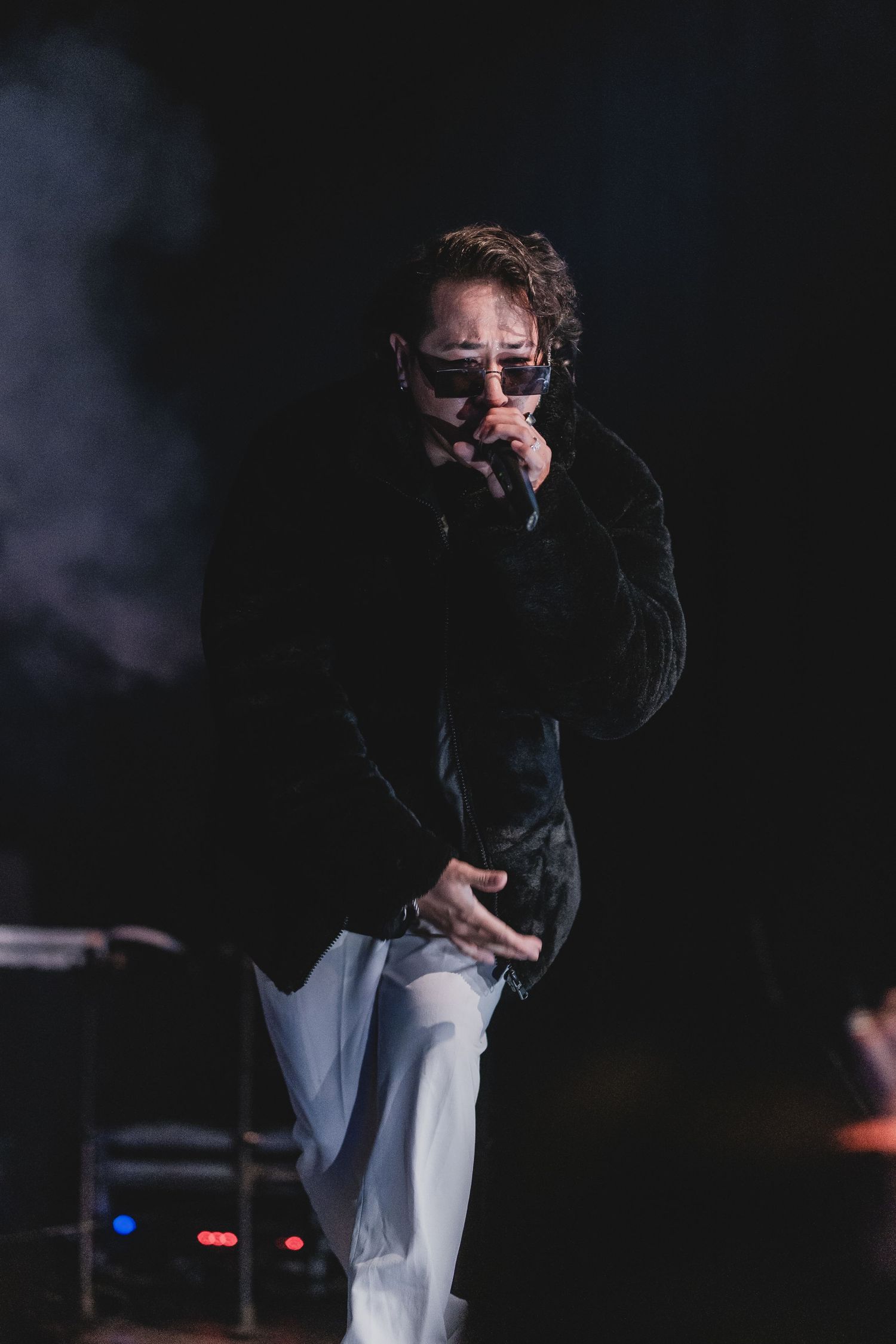 Performer in dark clothing singing on dimly lit stage with atmospheric smoke effects.