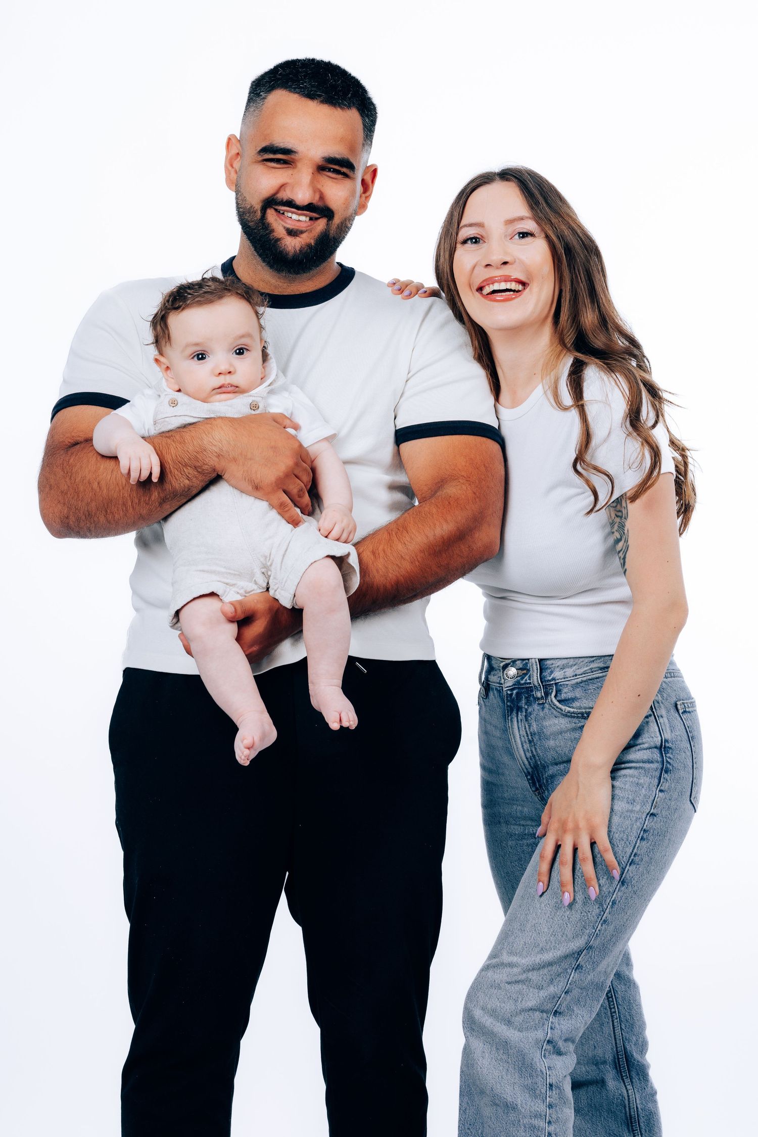 Family wearing matching white t-shirts and jeans poses playfully against white background in studio portraits.