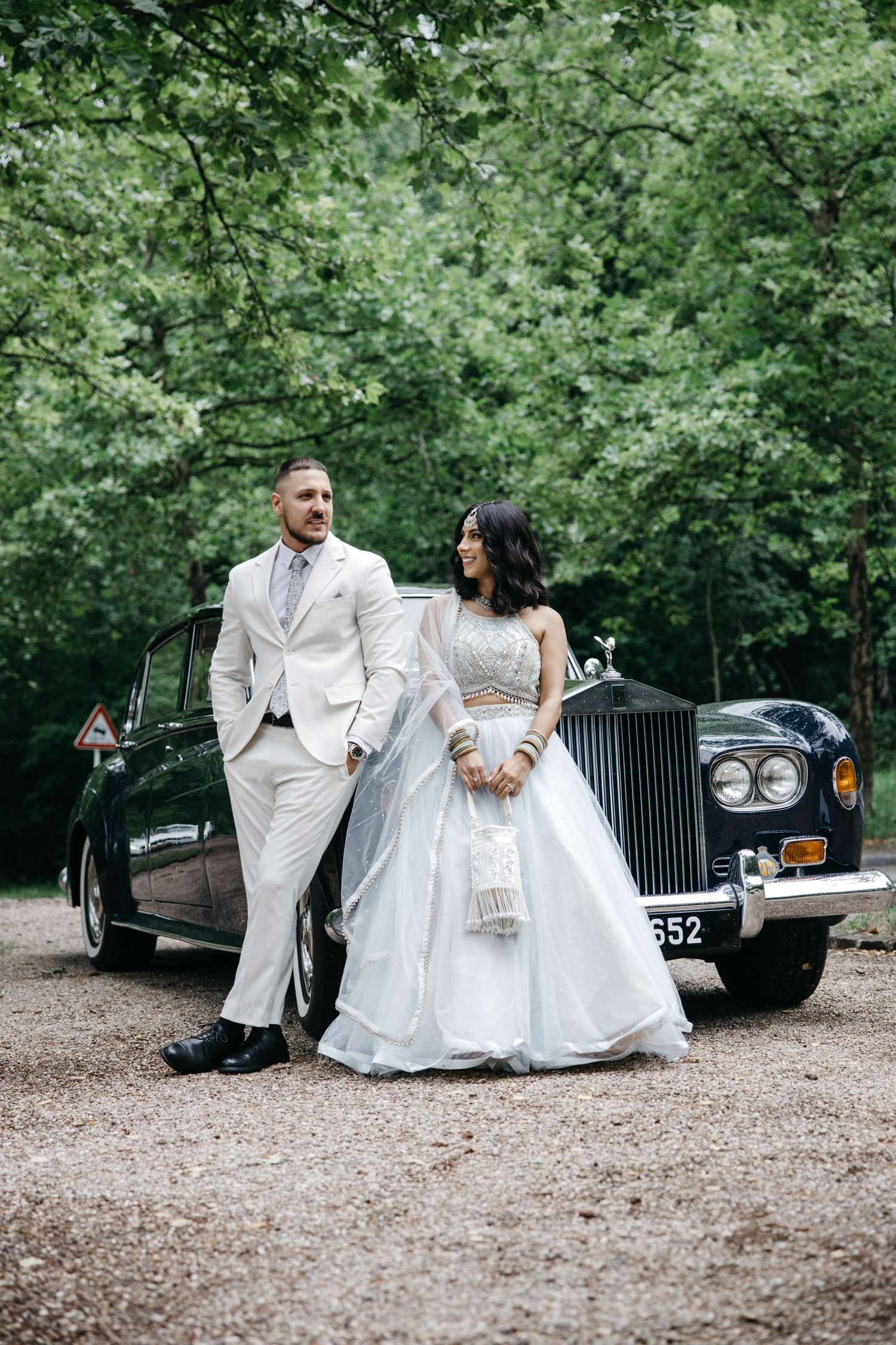 Elegant wedding couple posing near vintage black car in outdoor wooded setting.