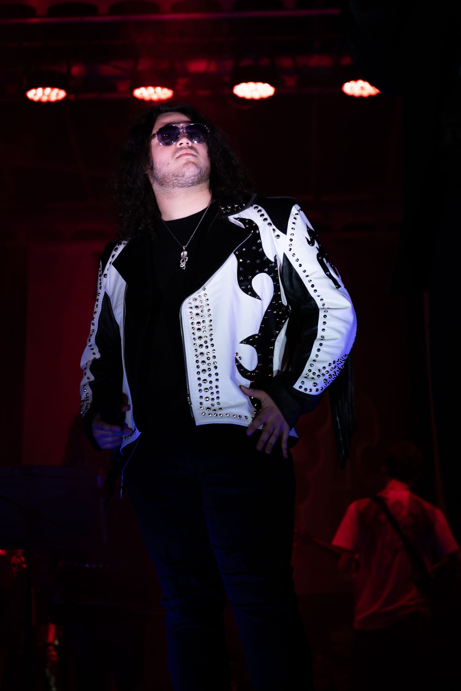 Performer in studded black and white jacket and sunglasses stands on stage under red and white lights.