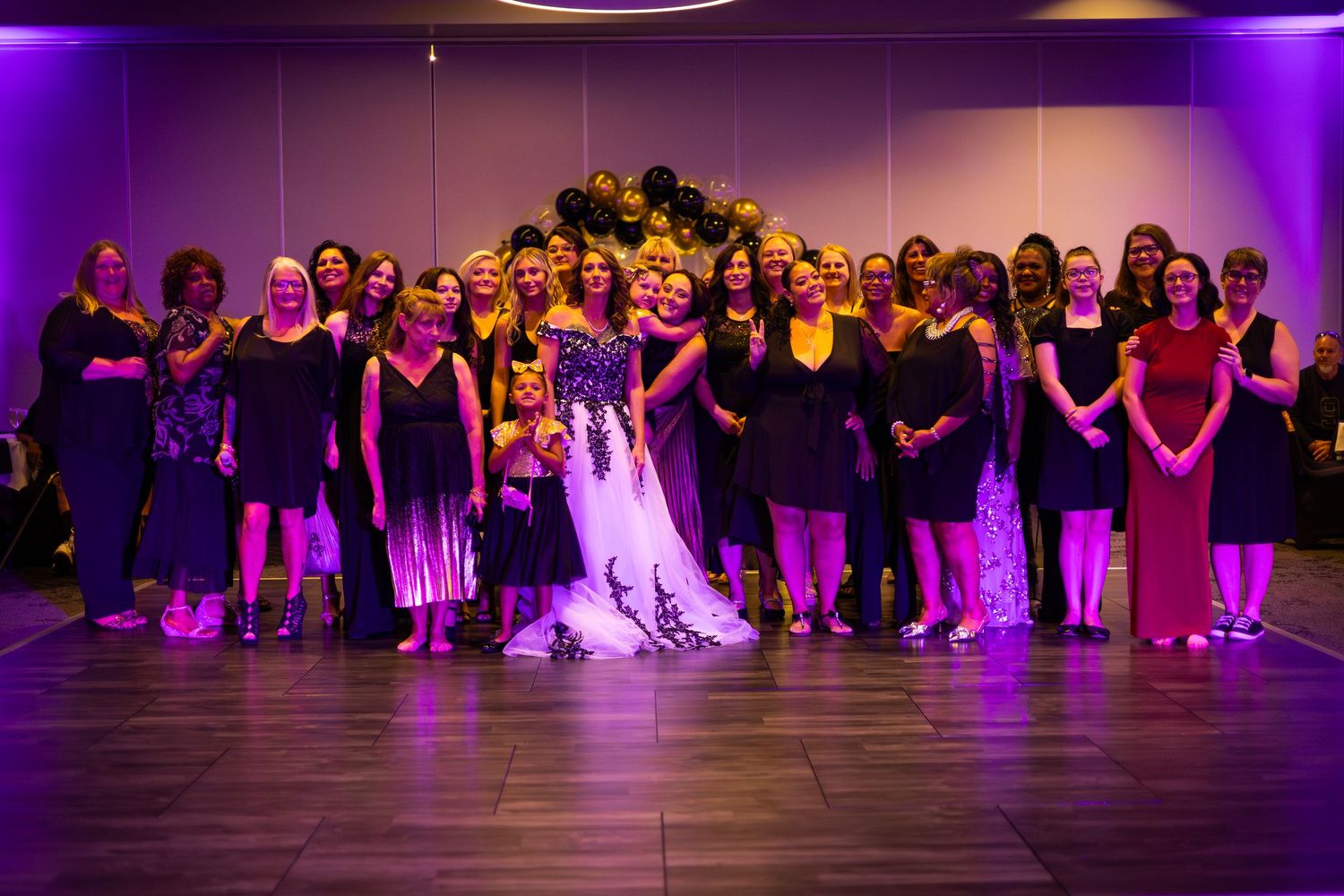 Large wedding party group photo with bride in center under purple lighting.