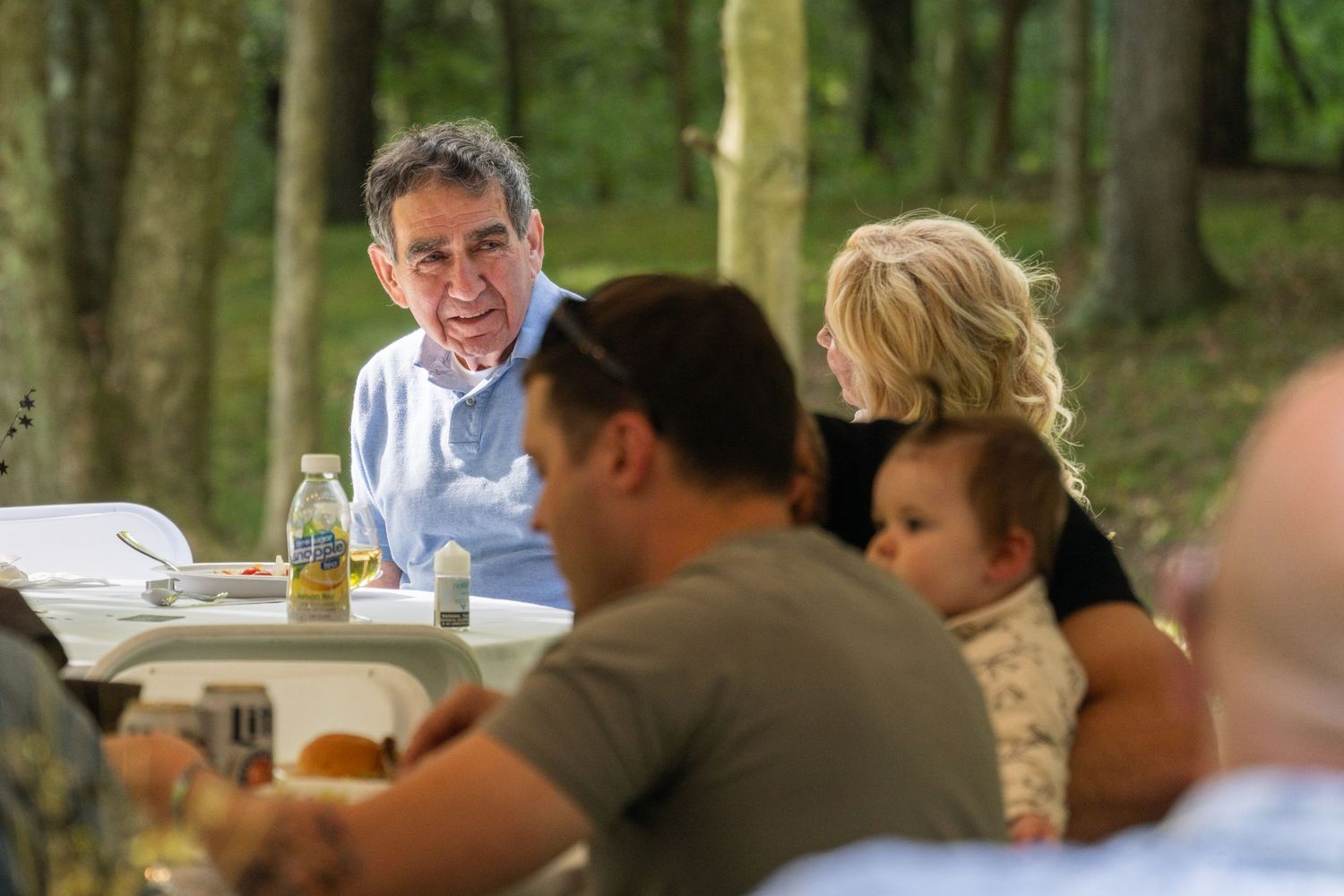 People seated at outdoor tables enjoying a casual gathering in a wooded setting.
