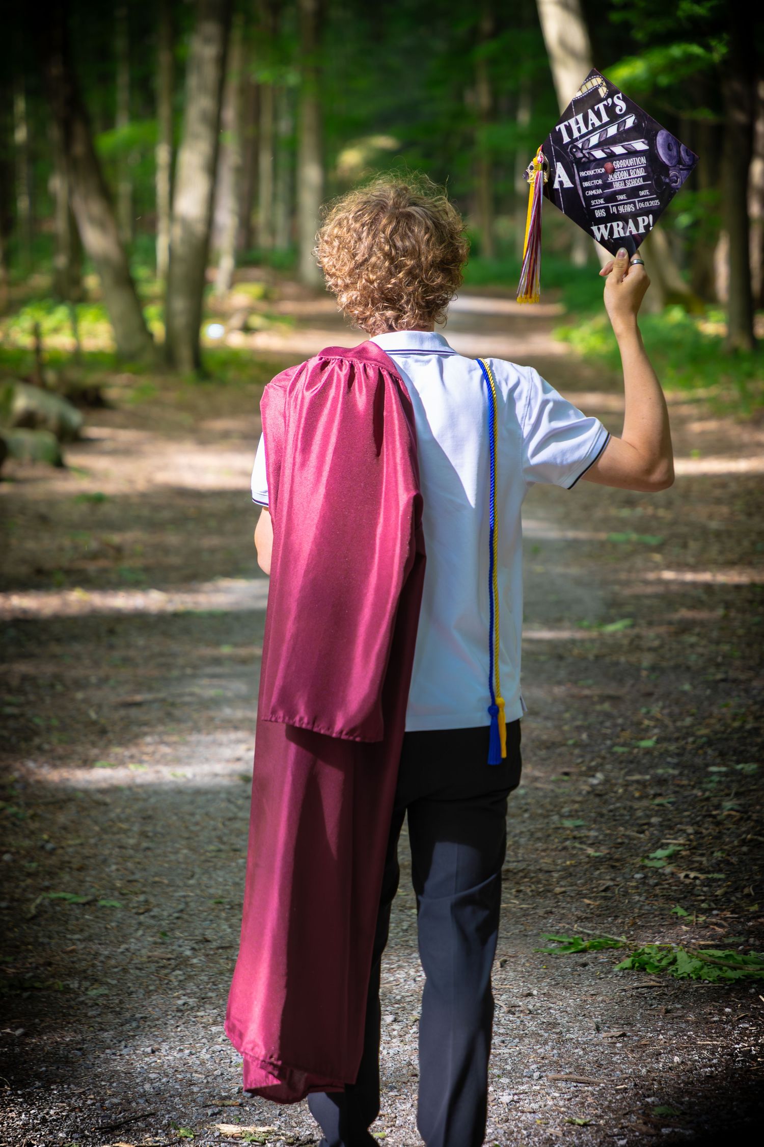 Two figures walking down a forest path carrying graduation items.