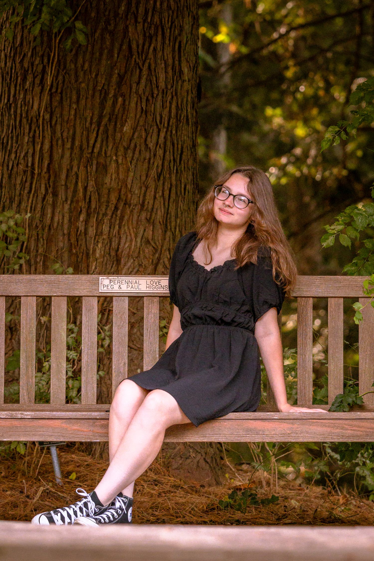 Person in black dress relaxing on wooden park bench surrounded by trees.