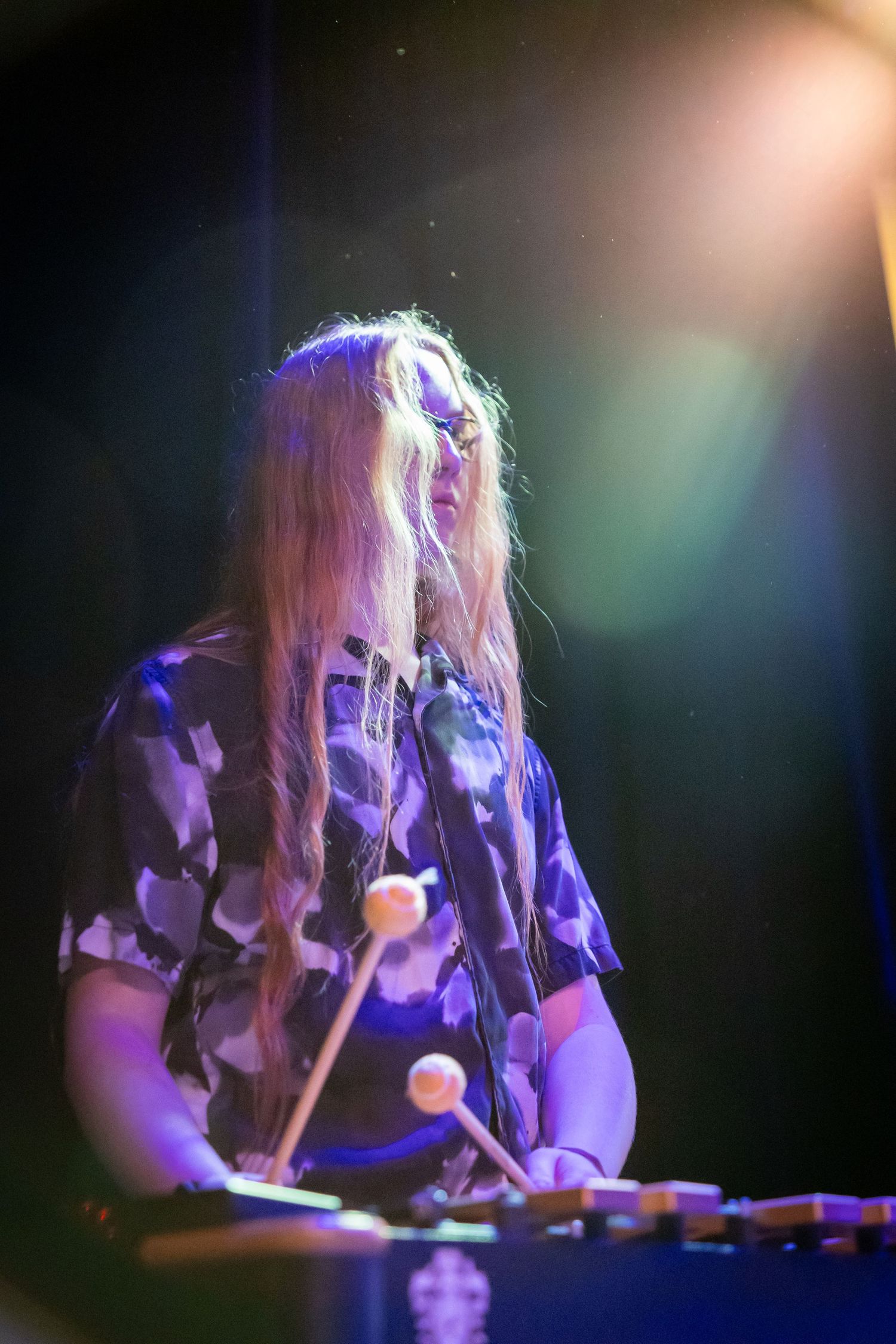 Long-haired musician playing marimba on stage under colorful purple and blue spotlights during live performance.