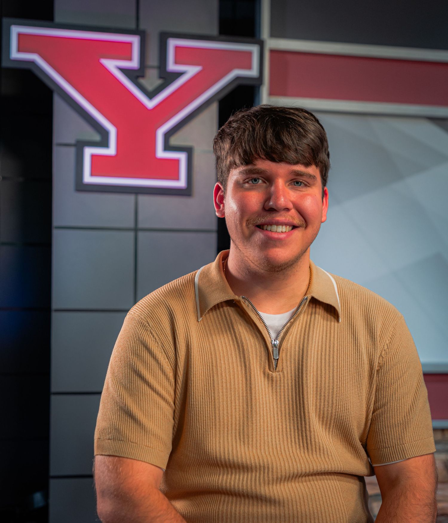 Young man in tan zip polo shirt smiling in front of a large red and white Y logo at Youngstown State University.