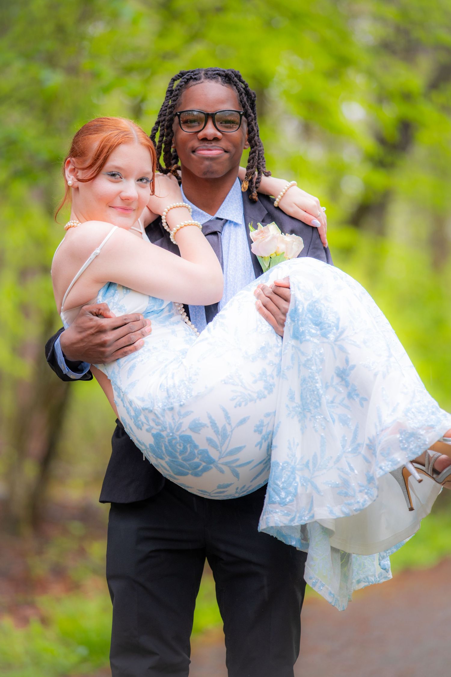 Groom carrying bride in floral dress outdoors in green forest setting, interracial couple in formal wedding attire.
