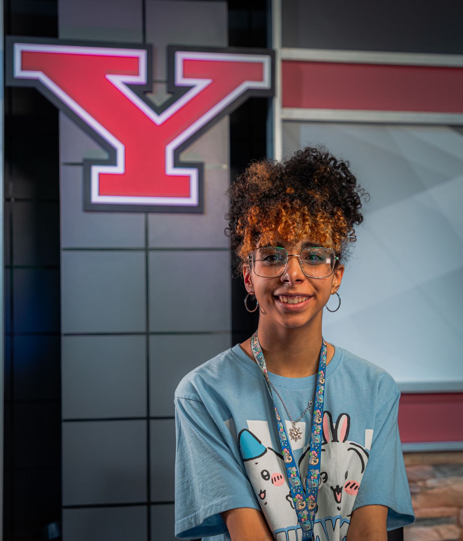 Smiling young woman with curly hair and glasses wearing a blue graphic tee stands in front of a red Youngstown State University Y logo.