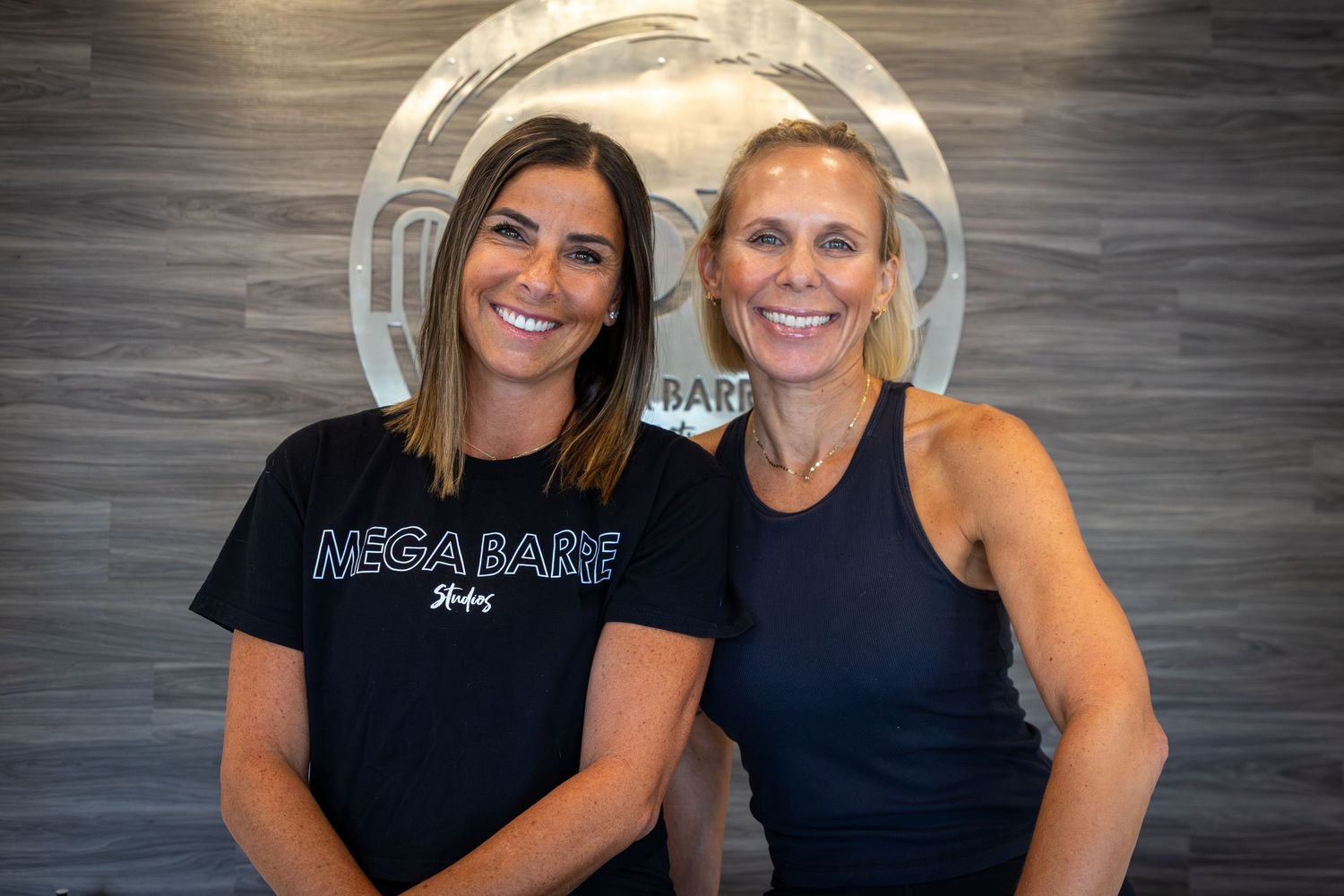 Two fitness instructors posing together in a studio with branded wall decor.