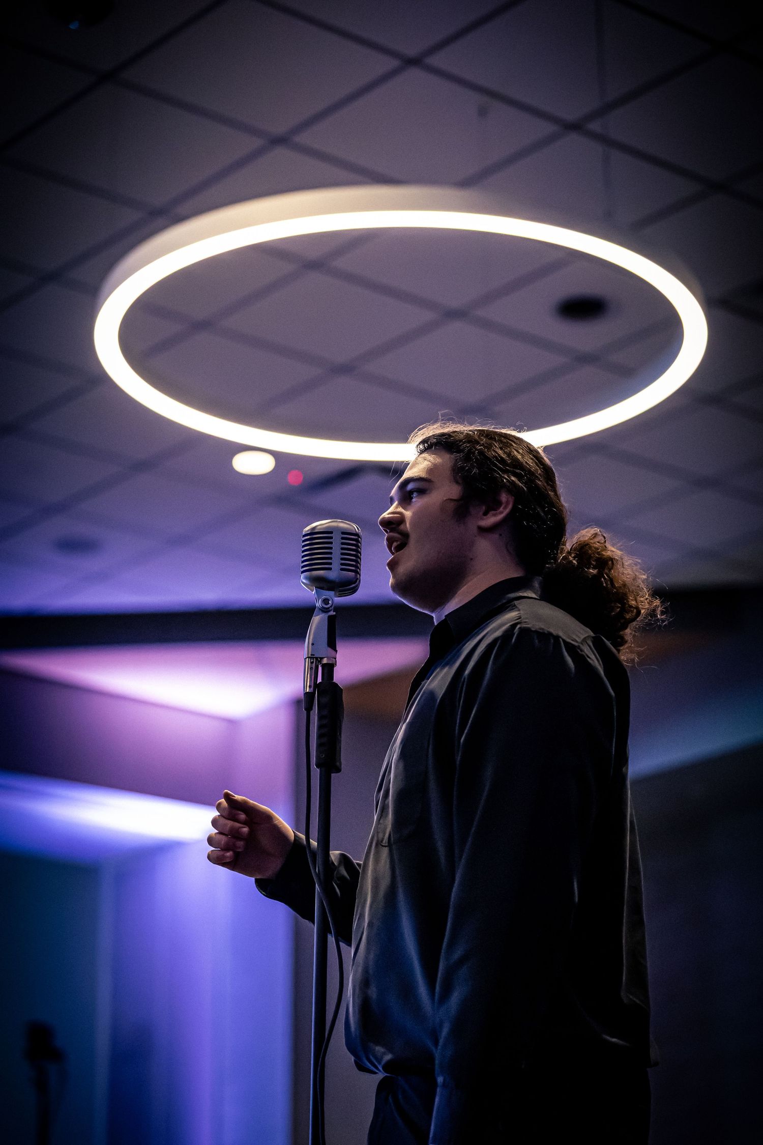Man singing passionately at a vintage microphone stand under a circular neon light in a dark blue-lit venue.