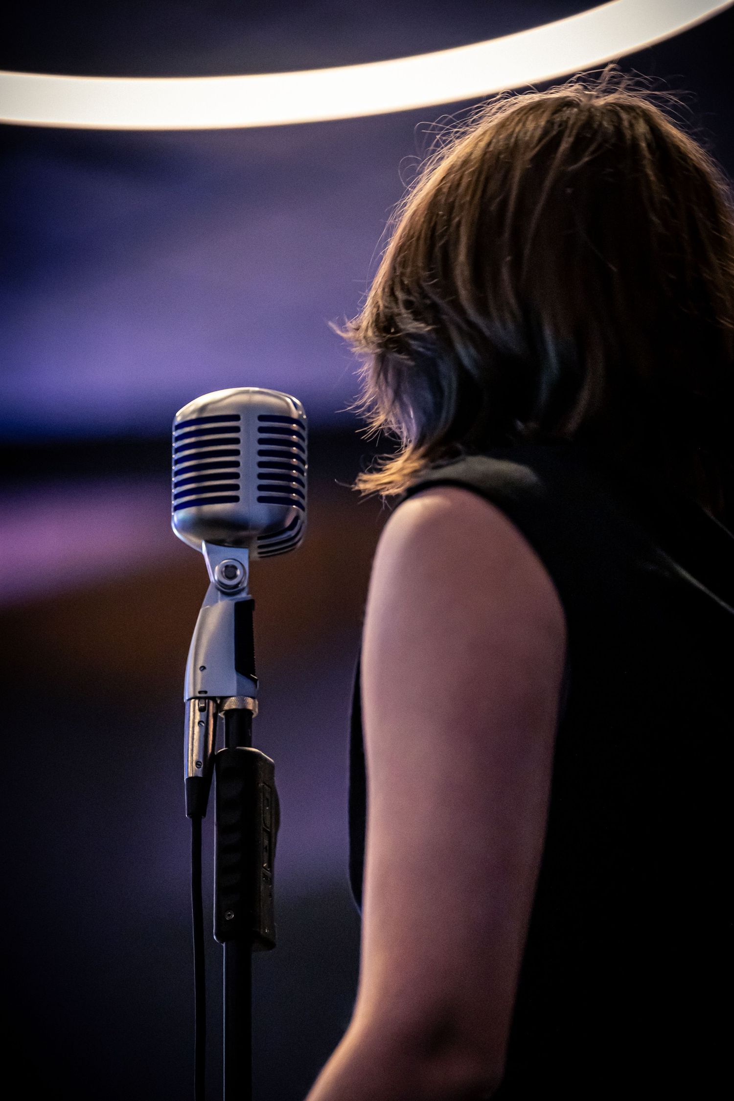 A female vocalist performing at a vintage silver microphone on stage under dramatic purple and blue stage lighting.