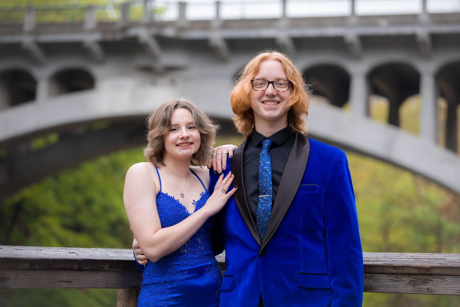 Two teens in matching royal blue formal wear pose smiling before a stone bridge for prom photos.