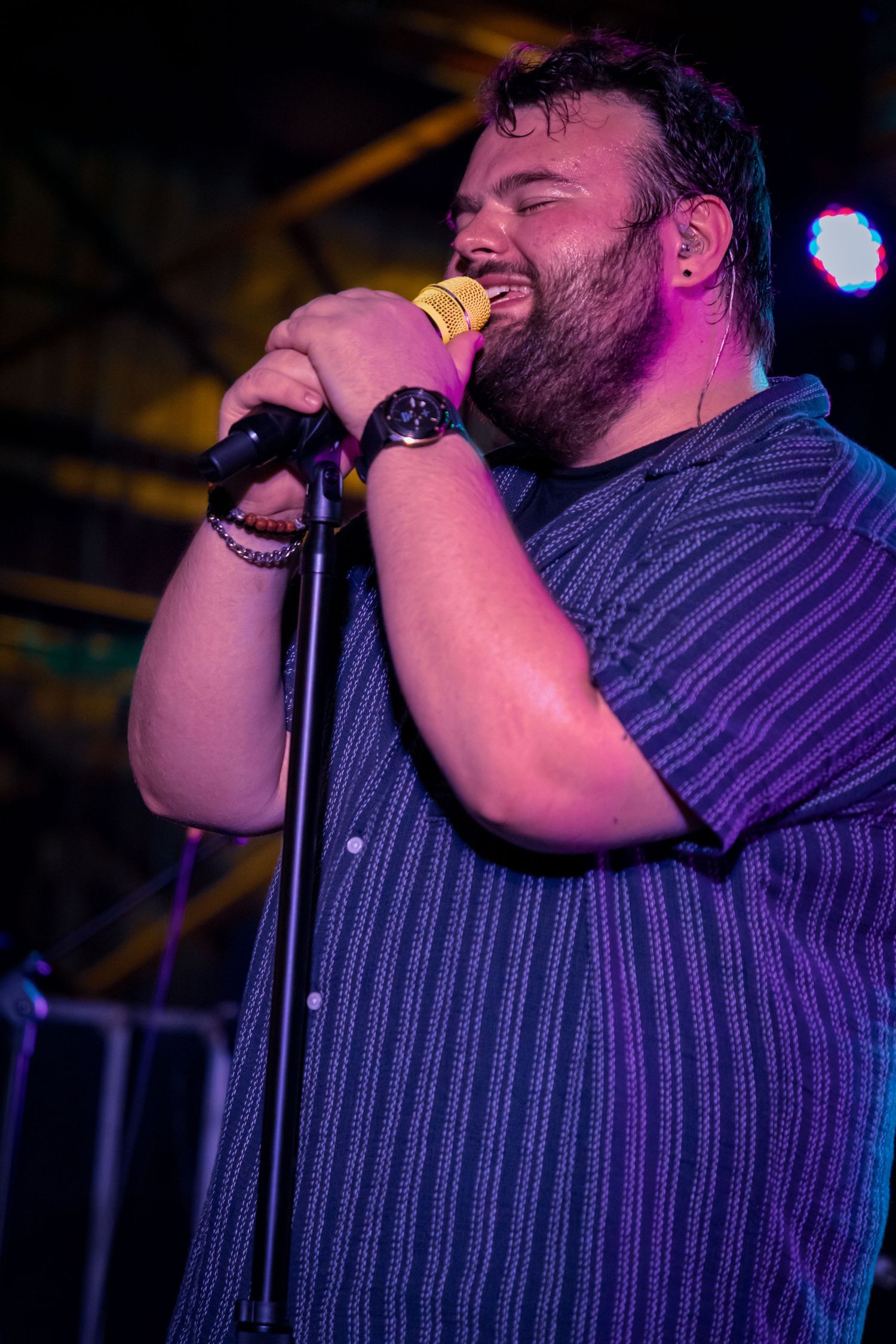 Singer performing passionately into microphone wearing striped shirt under purple stage lighting.
