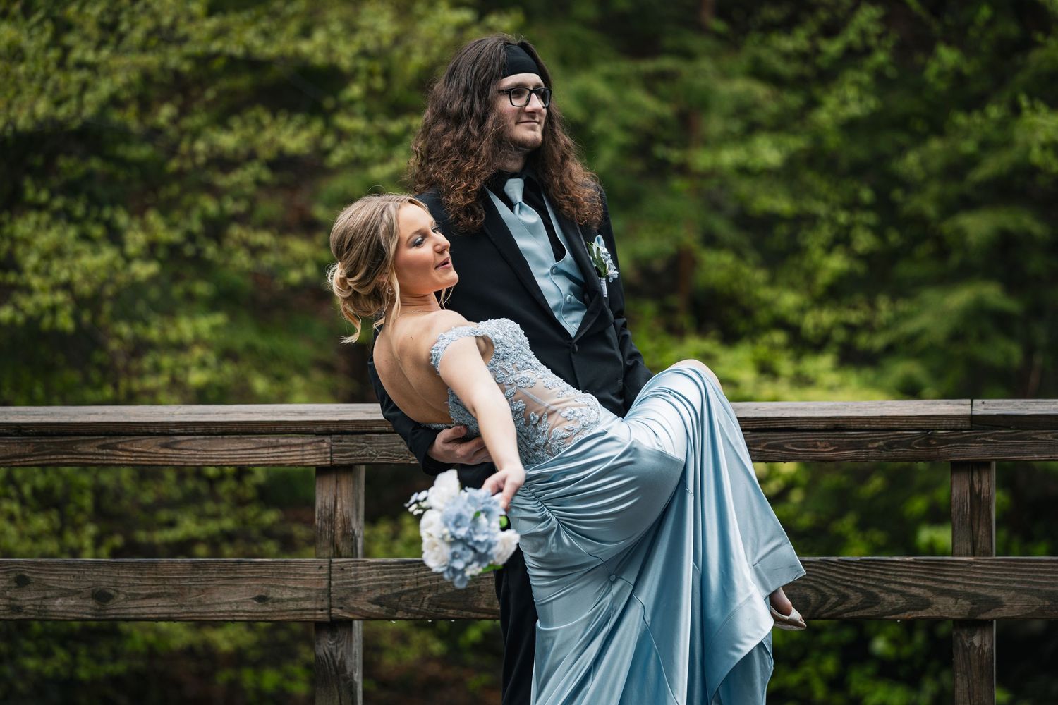 Couple in formal attire posing on a wooden bridge, woman in blue gown holding bouquet, man in black tuxedo, forest backdrop.