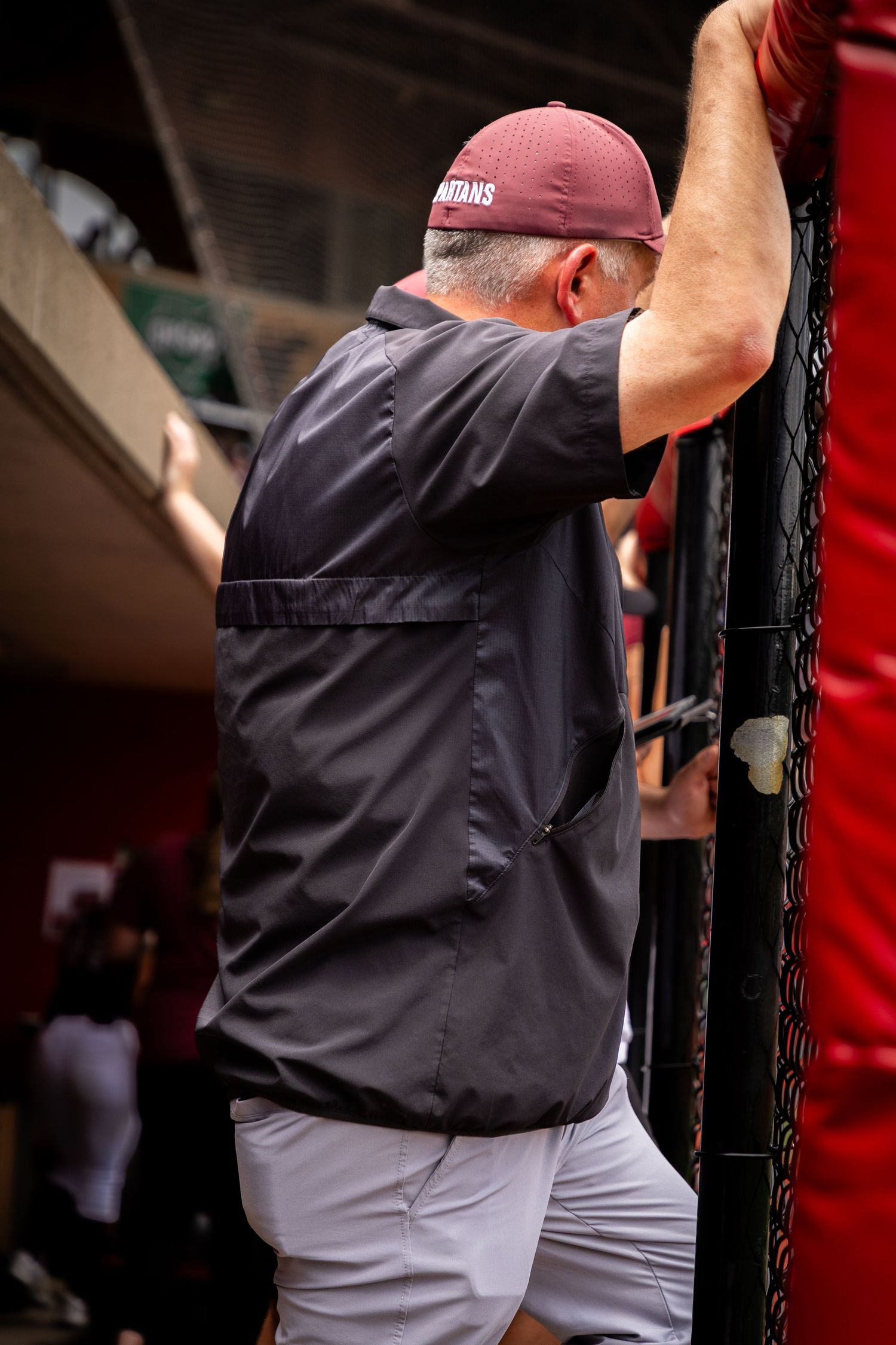 Baseball coach watches game from dugout wearing navy shirt and grey pants.