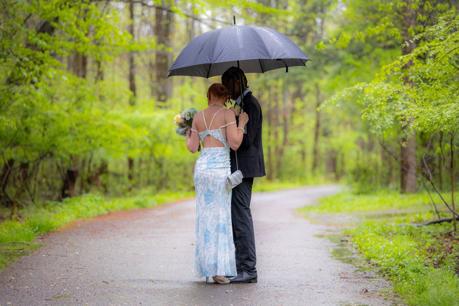 Couple sharing umbrella on rainy forest path, woman in blue gown holding bouquet, man in black suit.