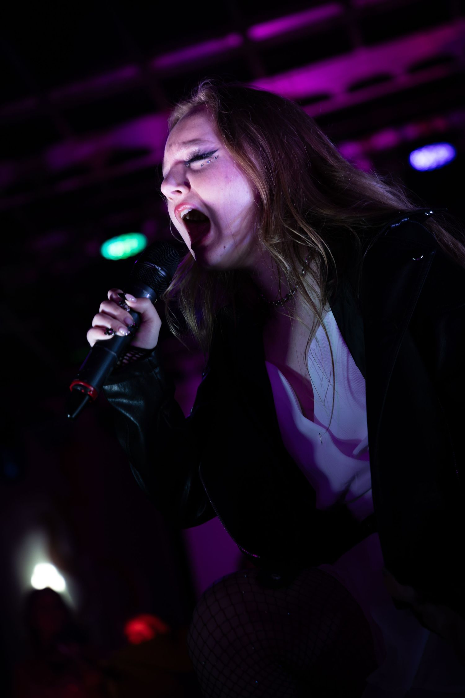 Female vocalist singing passionately into microphone on stage under purple lights wearing black leather jacket.