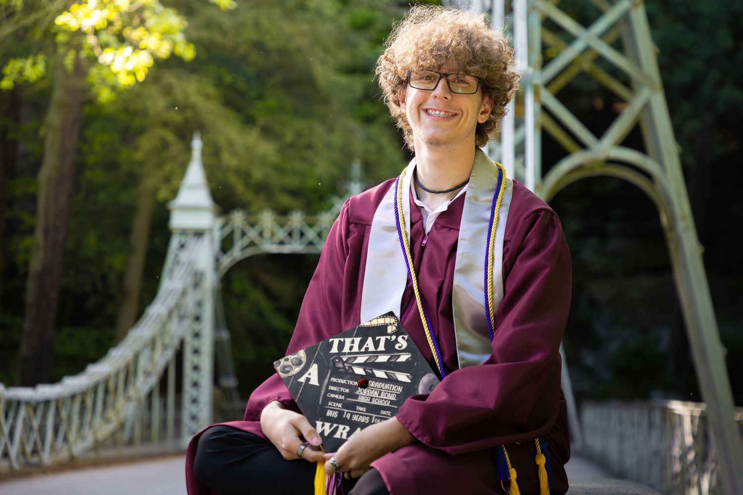 Graduate in maroon cap and gown sits on bench near suspension bridge in outdoor setting.