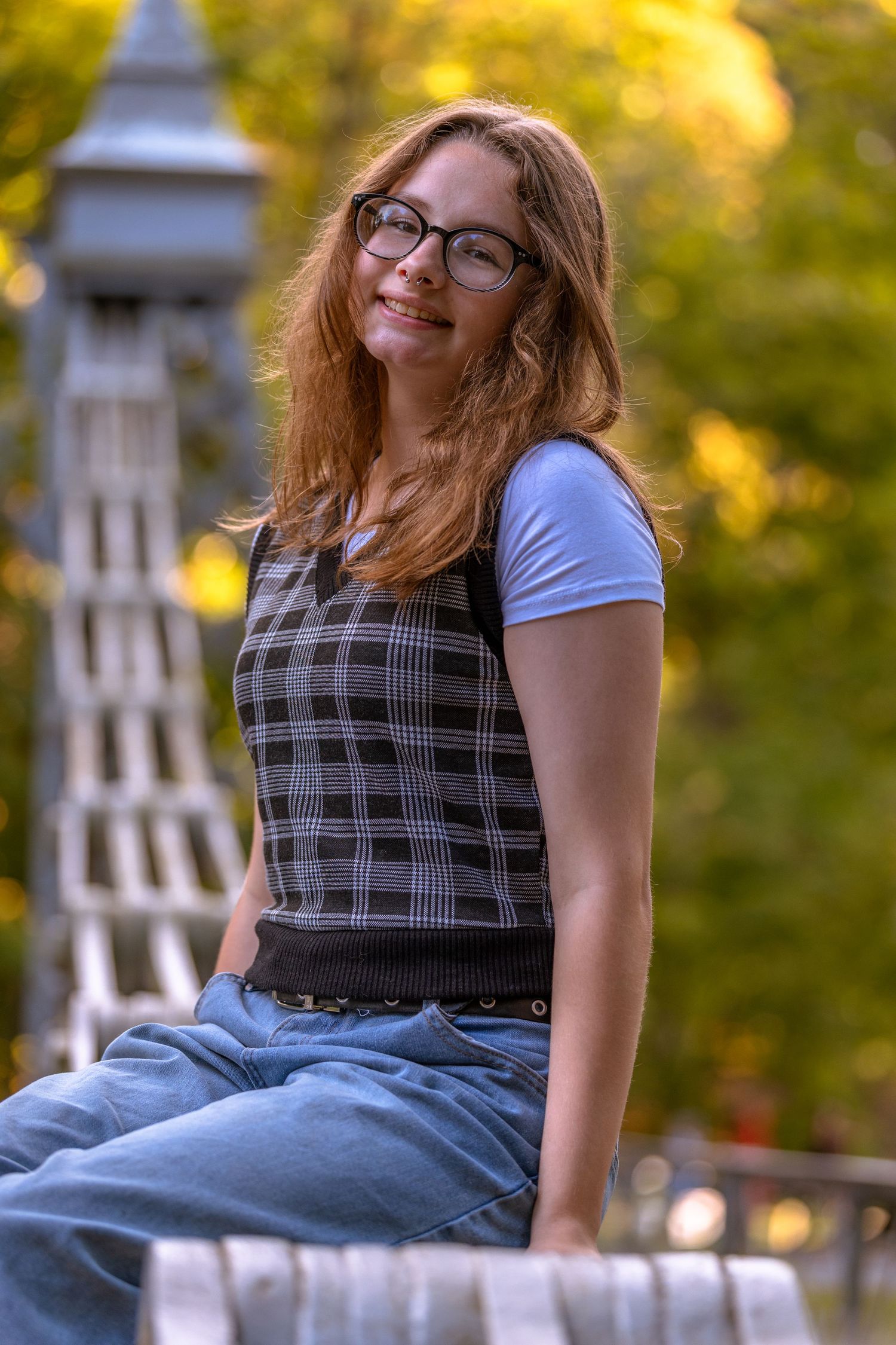 Person in plaid dress and glasses sits on white railing with autumn foliage in background.