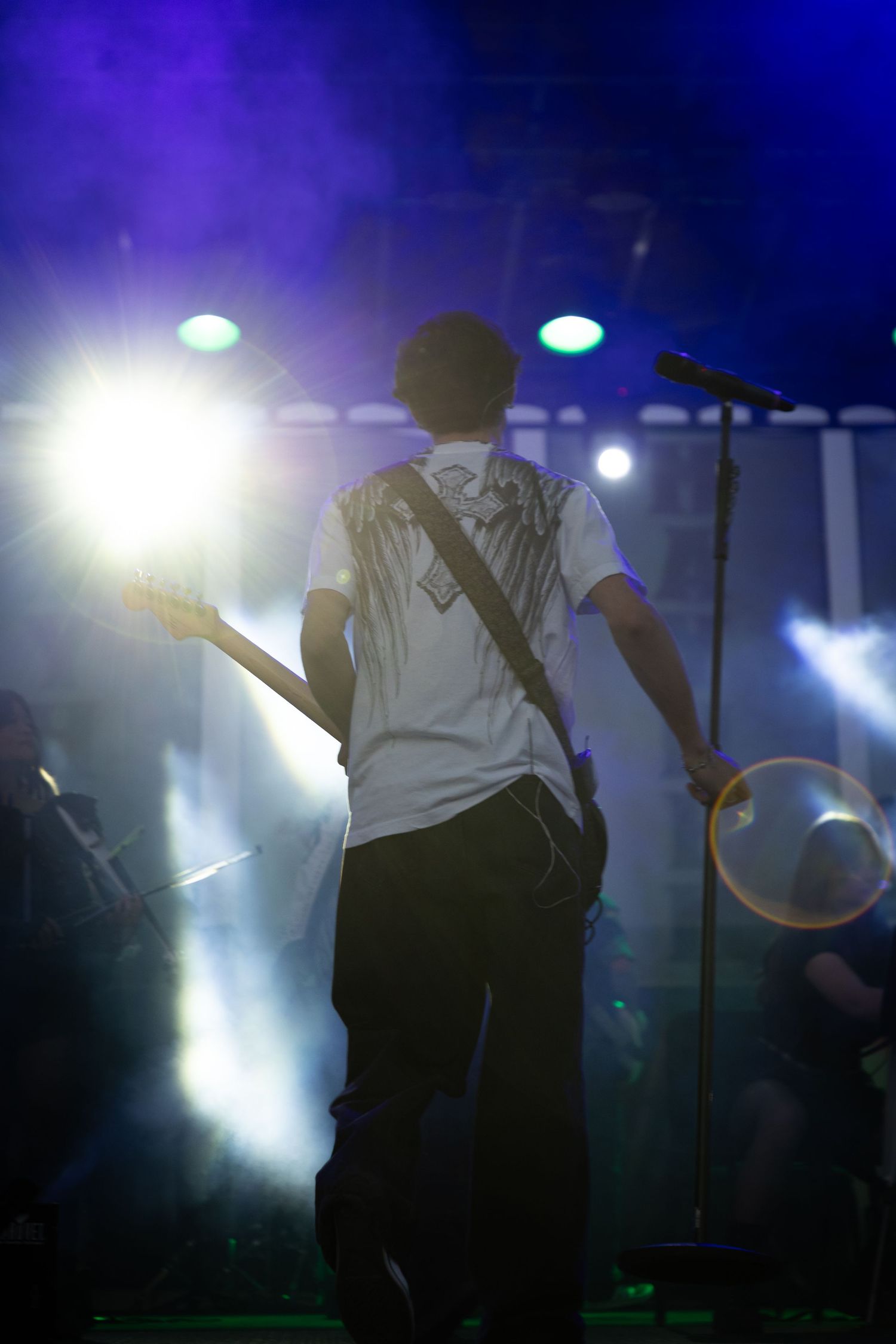 Guitarist performing on stage from behind, wearing white t-shirt, with dramatic blue and green concert lighting.