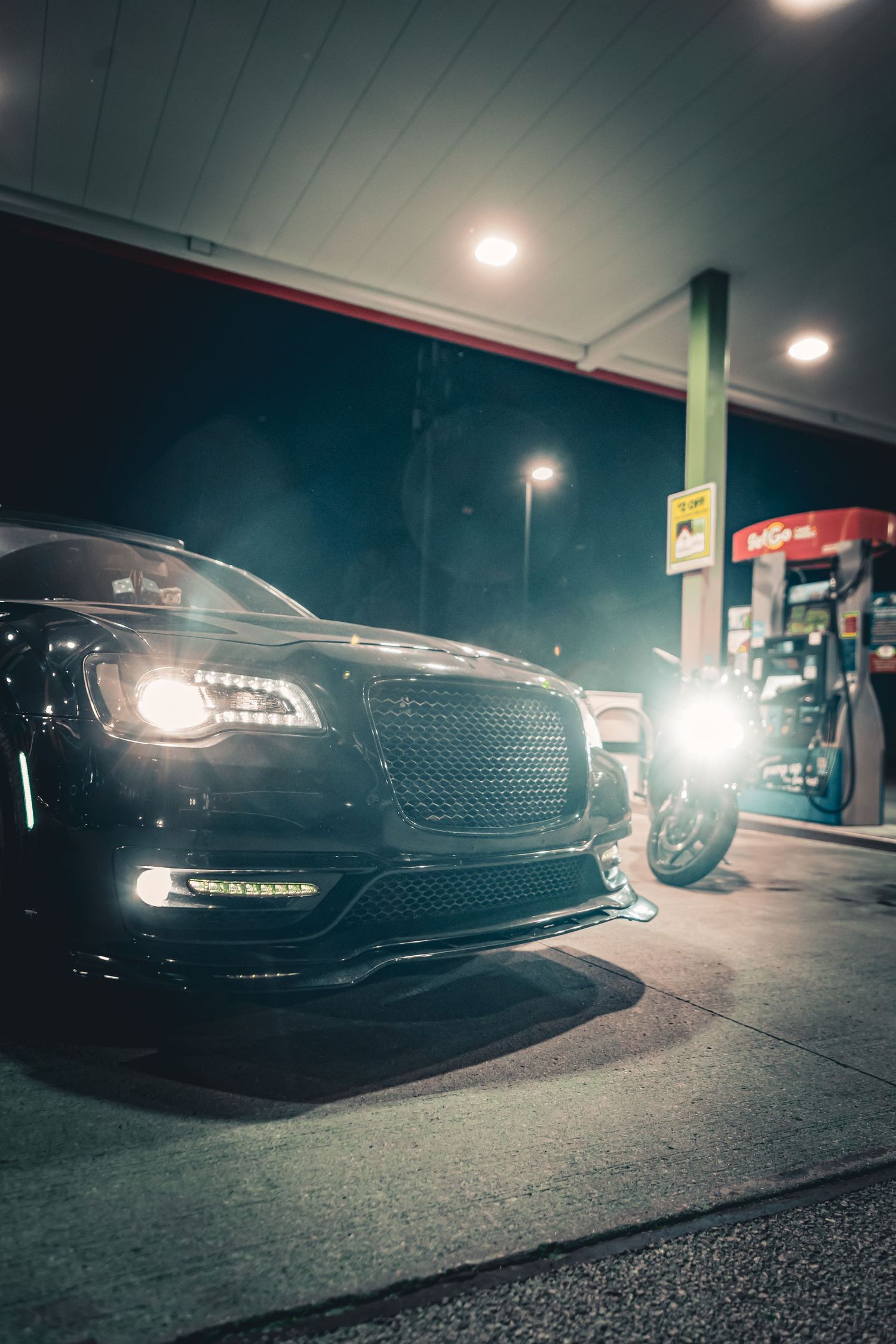 Black luxury sedan and motorcycle parked at a gas station at night with bright lights illuminating the scene.