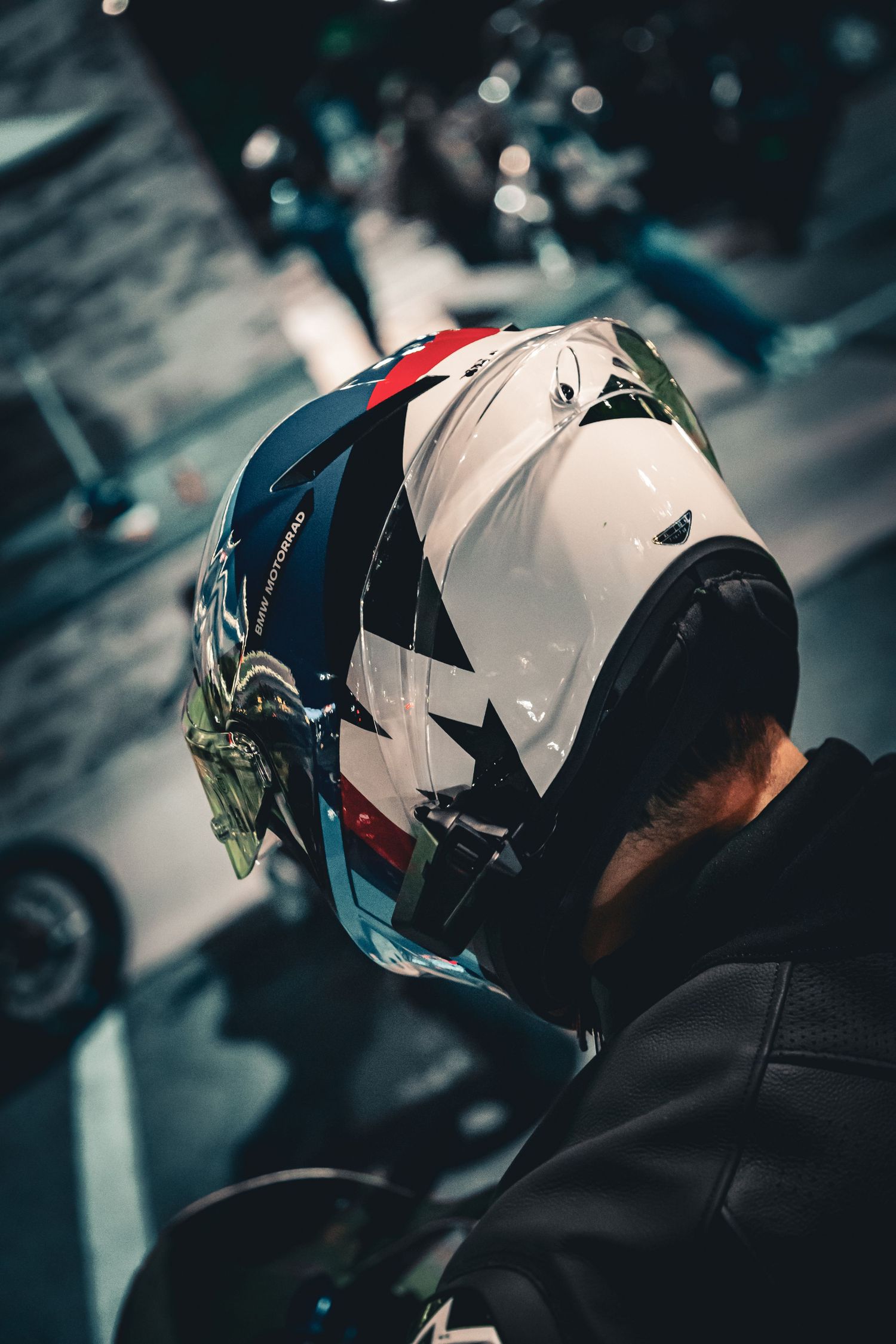 Motorcyclist wearing a white helmet with star design and red, blue accents, shot from behind in moody dark setting.