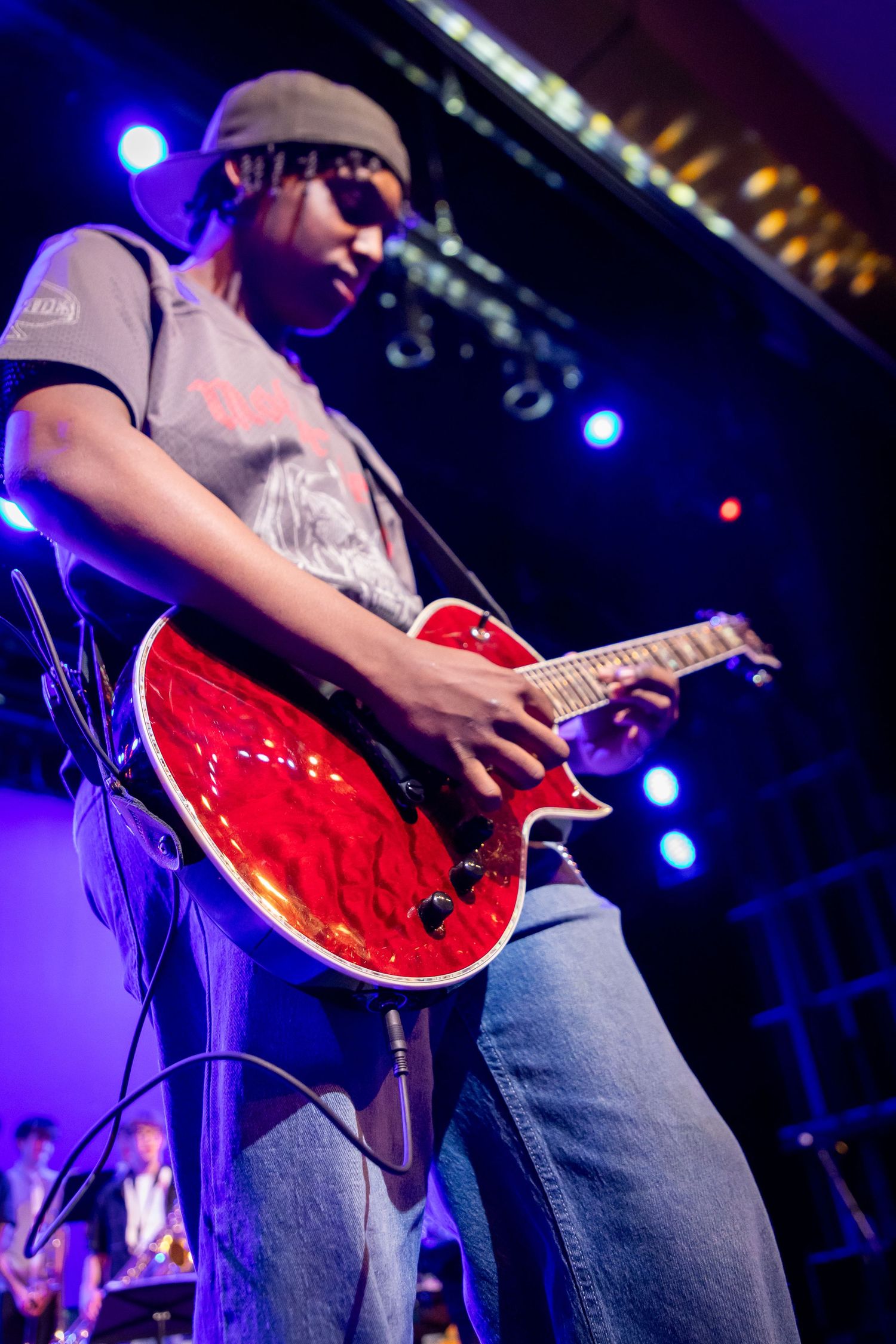 Young musician playing red electric guitar on stage under blue and purple lights during a live performance.