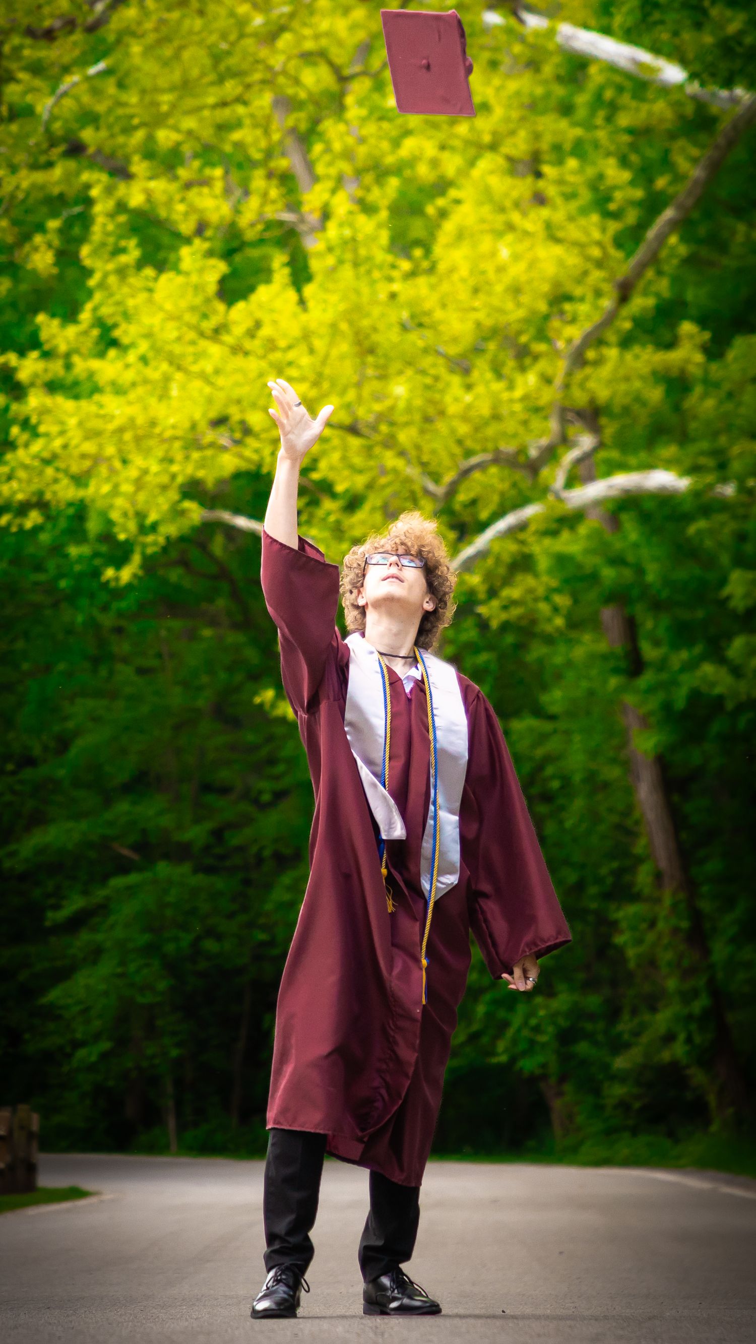 Graduate in maroon gown throws diploma in air against vibrant green foliage background.
