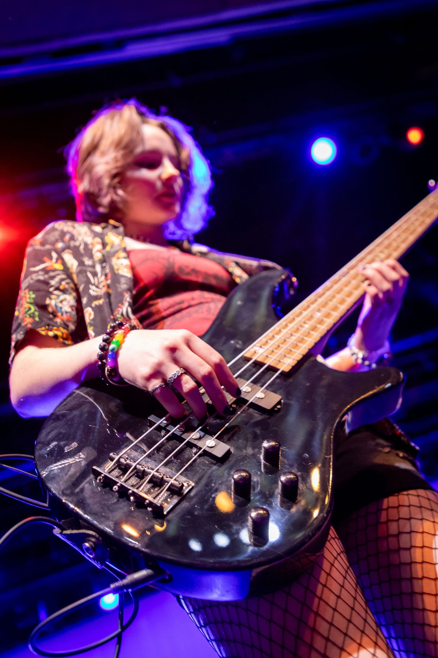 Female bassist performing on stage under colorful lights, playing a worn black bass guitar while wearing fishnet stockings.