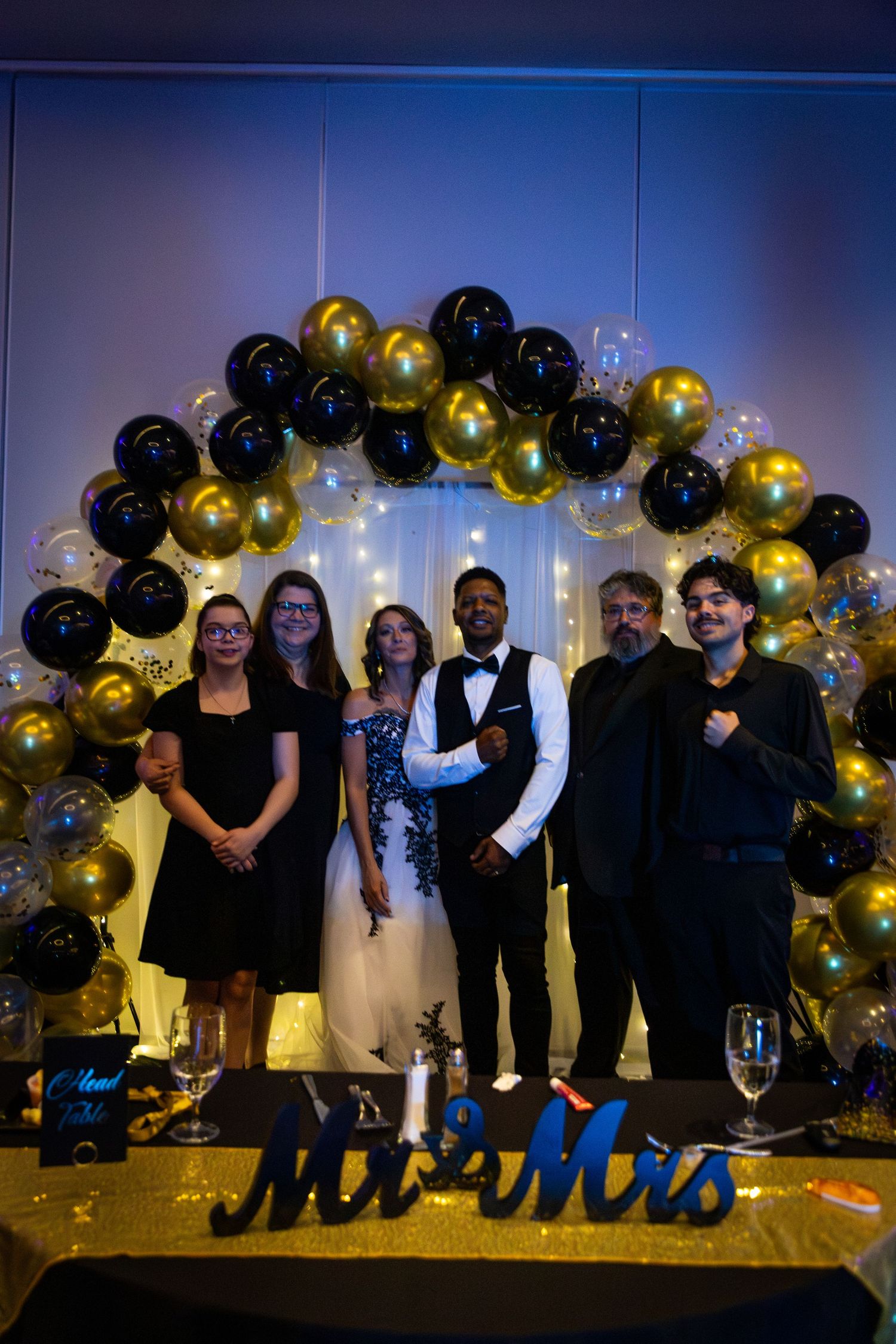 Wedding party poses under elegant black and gold balloon arch at reception.