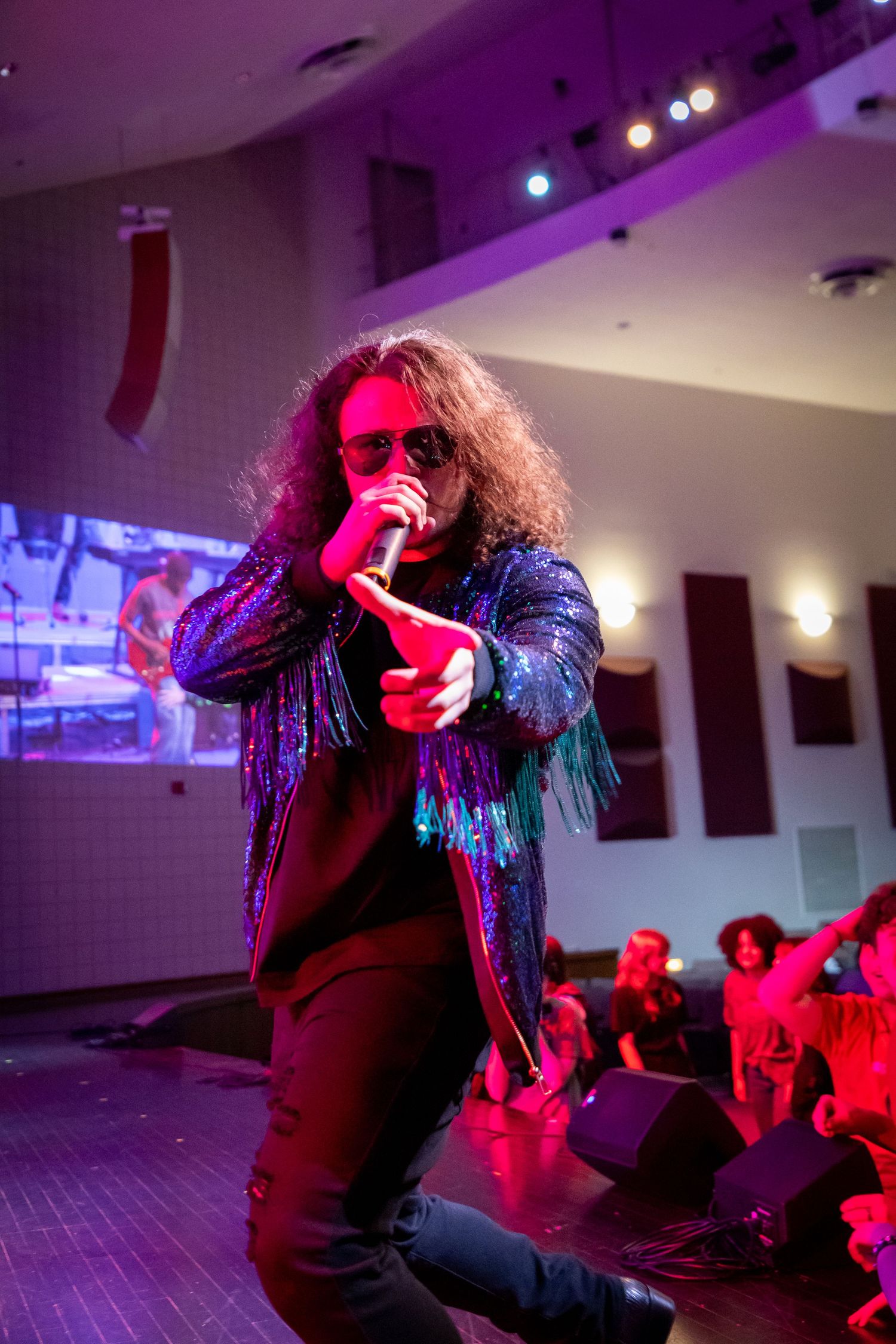 A rock singer with long hair performs on stage wearing a sequined jacket under purple lights at an indoor concert venue.
